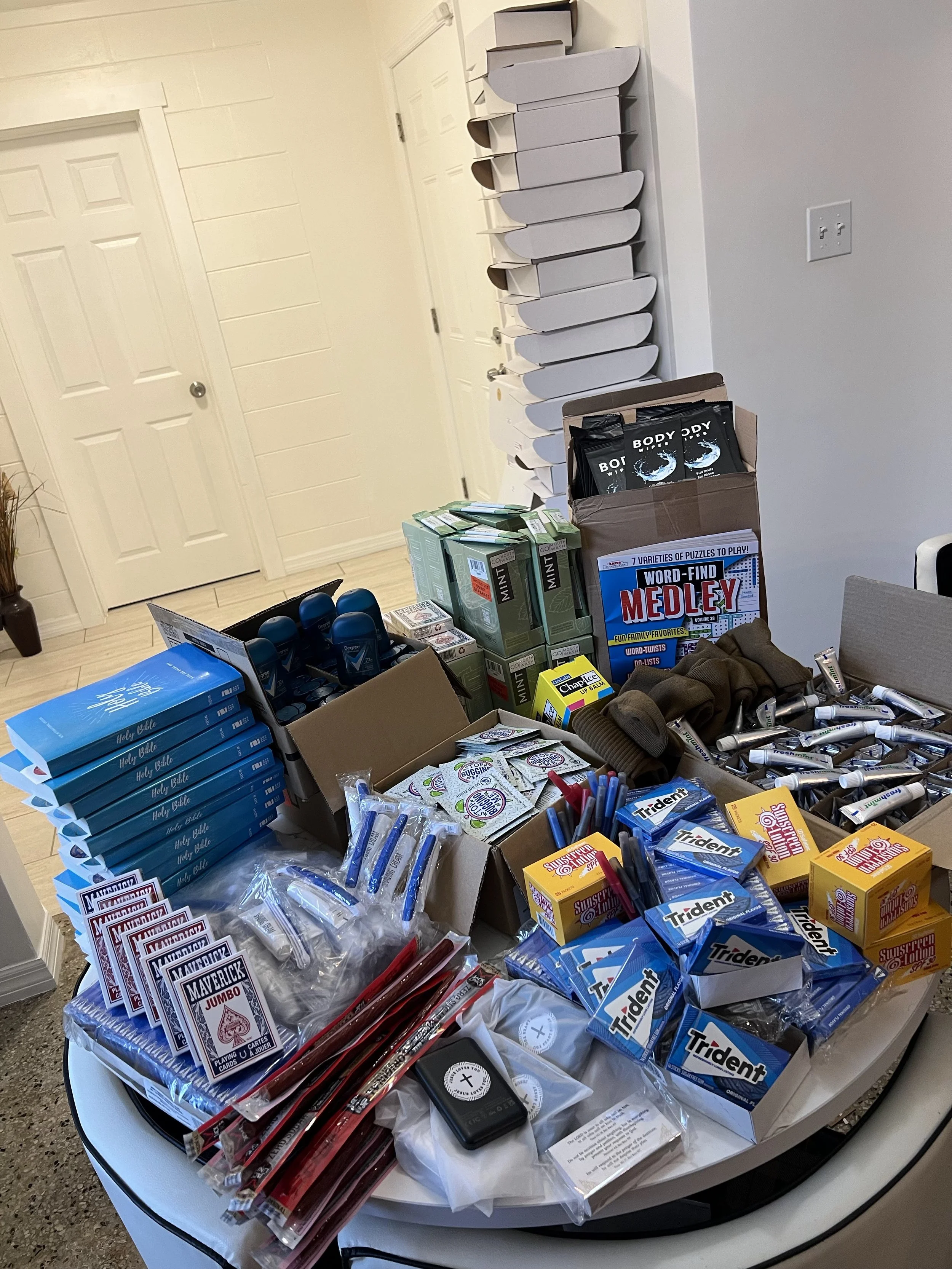 Assorted items including boxes of Holy Bibles, a stack of playing cards, tubes of toothpaste, packs of gum, challenge coins, word-find books, and other miscellaneous items organized on a table. There are also empty boxes stacked against the wall.