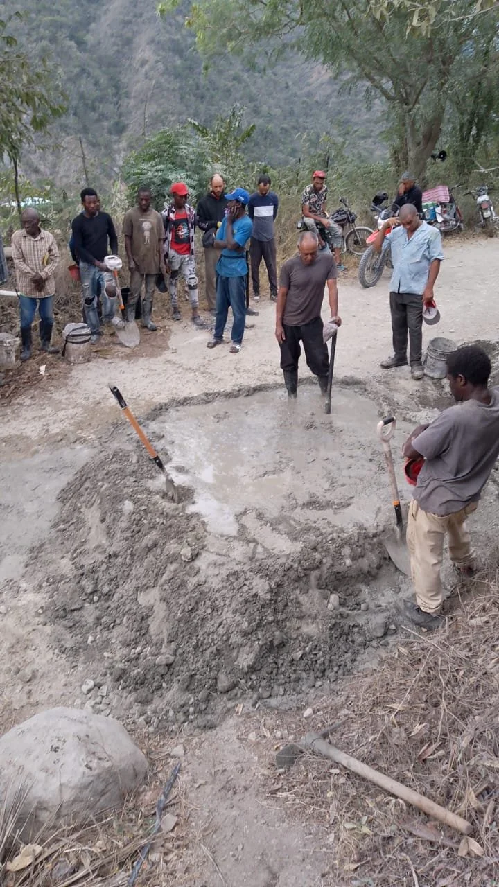 Group of people mixing cement with shovels near a dirt path; motorcycles and trees in background.