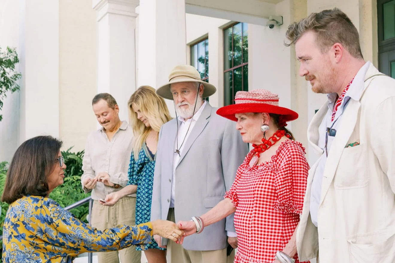 A group of six diverse people, including an older woman in red gingham and red hat, shaking hands with a woman in glasses and a colorful shirt, outside in front of a building with large windows.