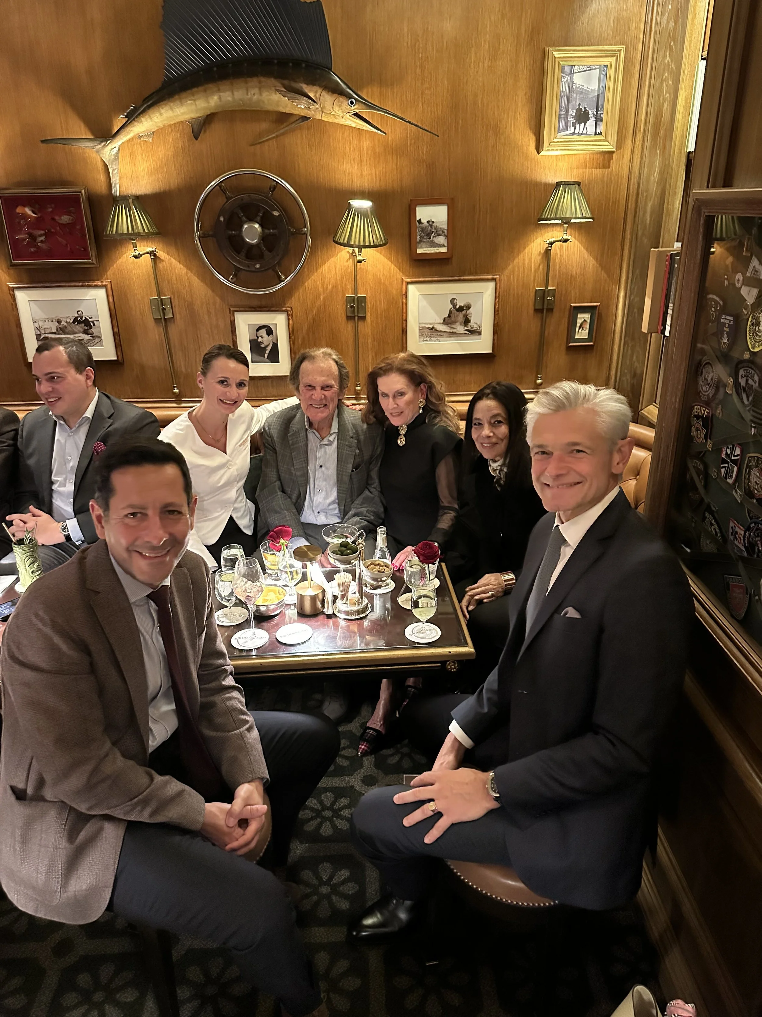 Group of people smiling and sitting around a dinner table in a restaurant with wooden walls adorned with framed photographs, a mounted fish, ship wheel, and wall lamps.