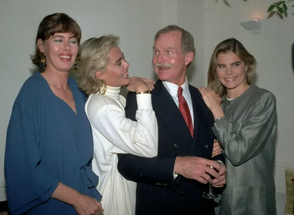 Four people, three women and one man, are smiling and posing together at a social gathering indoors. The man is holding a glass of wine and the women are standing close to him, with one woman touching his shoulder and another resting her hand on his arm.