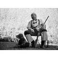 A person sitting on a park bench, reading a book and holding a walking stick, with a dog beside them.