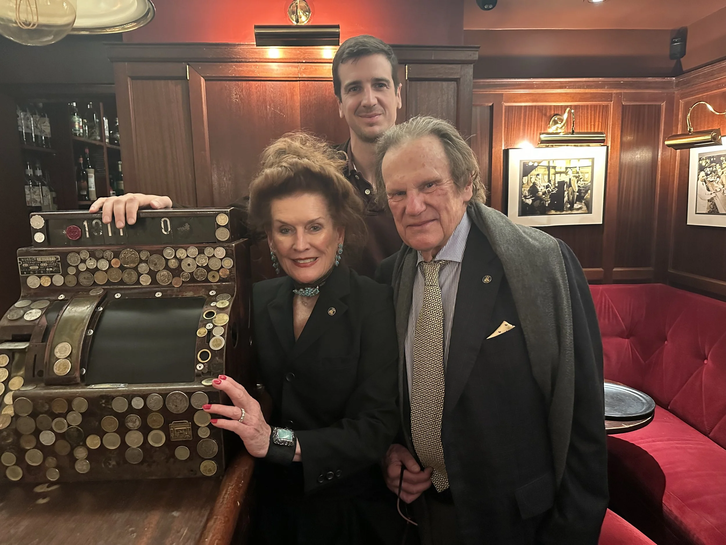 Three people in a bar or restaurant, standing behind a coin collection bank that is filled with coins, and smiling at the camera.