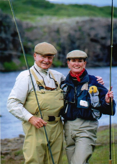 Two people in fishing gear smiling outdoors near water with rocky cliffs and green grass in the background, holding fishing rods.