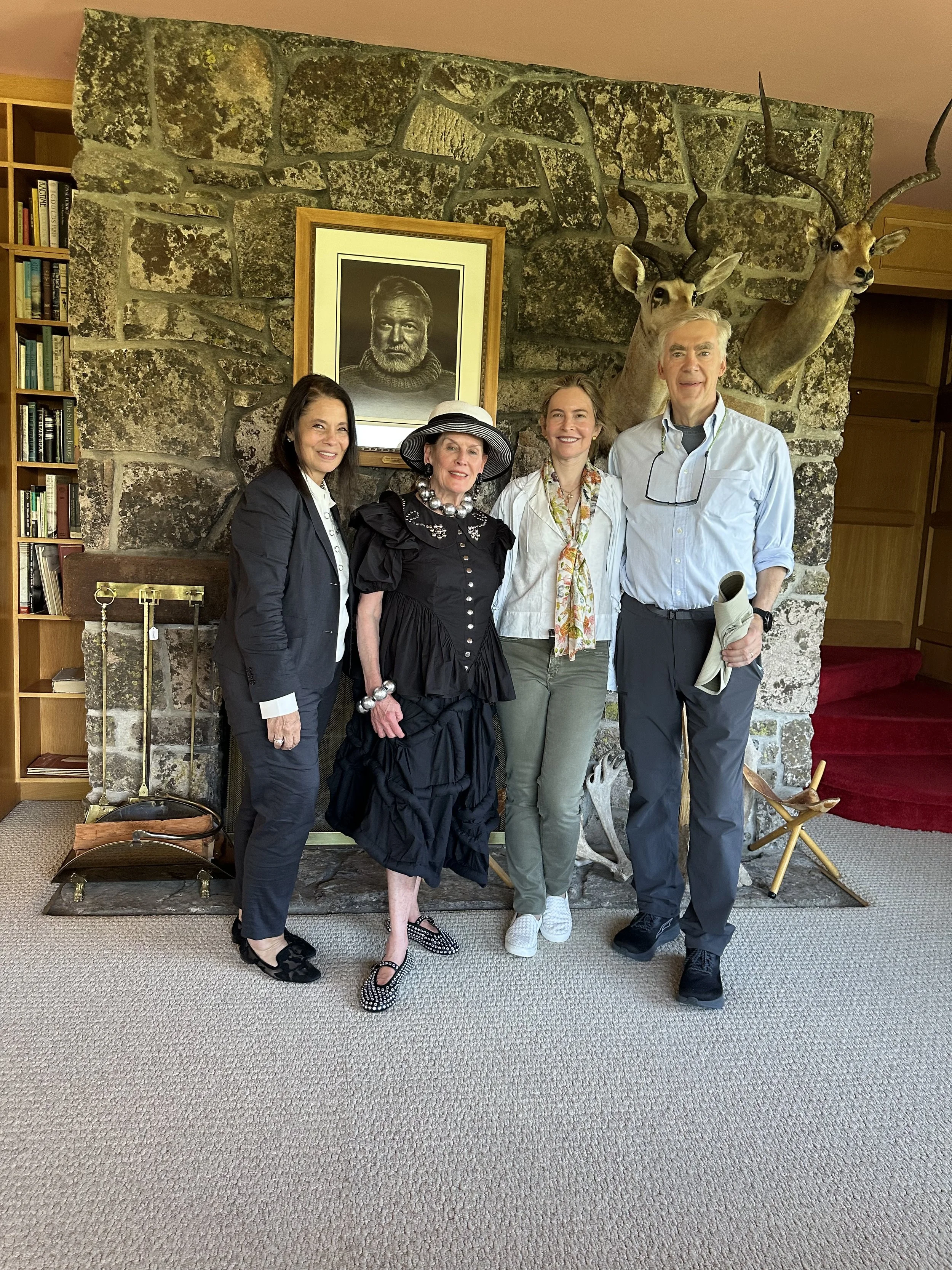 Four people standing in front of a stone fireplace with mounted animal heads and a portrait of a man, inside a room with bookshelves and a red velvet bench.