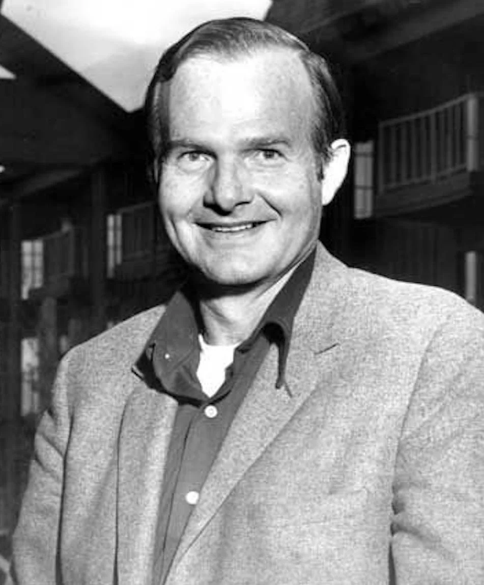 Black and white photo of a smiling man wearing a suit jacket over a shirt, standing indoors.