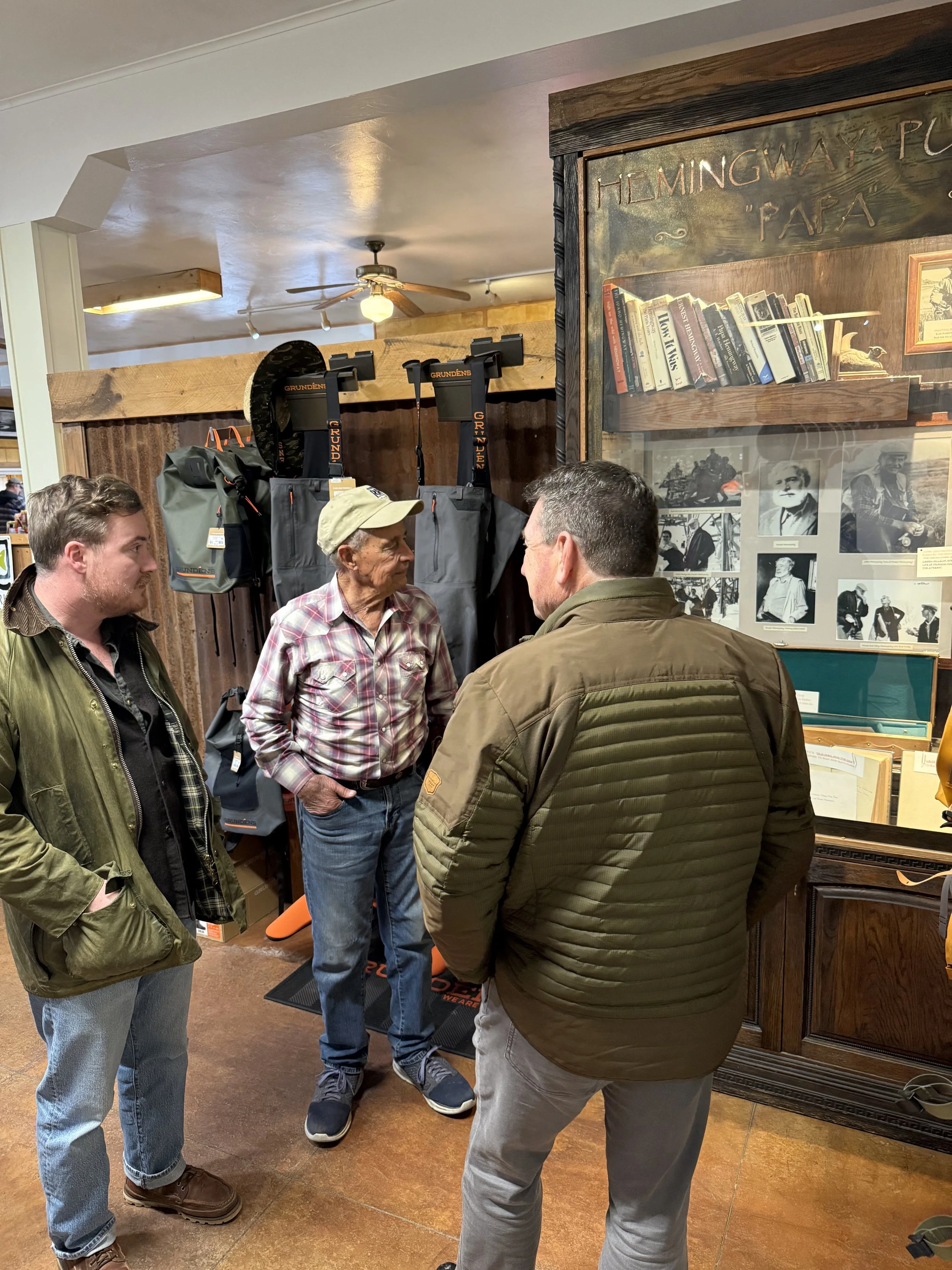 Four men engaged in conversation in a store with hunting and outdoor gear. One man is wearing a plaid shirt and baseball cap, another in a green jacket, and two others are dressed in dark jackets. The store has a wooden display with books and vintage