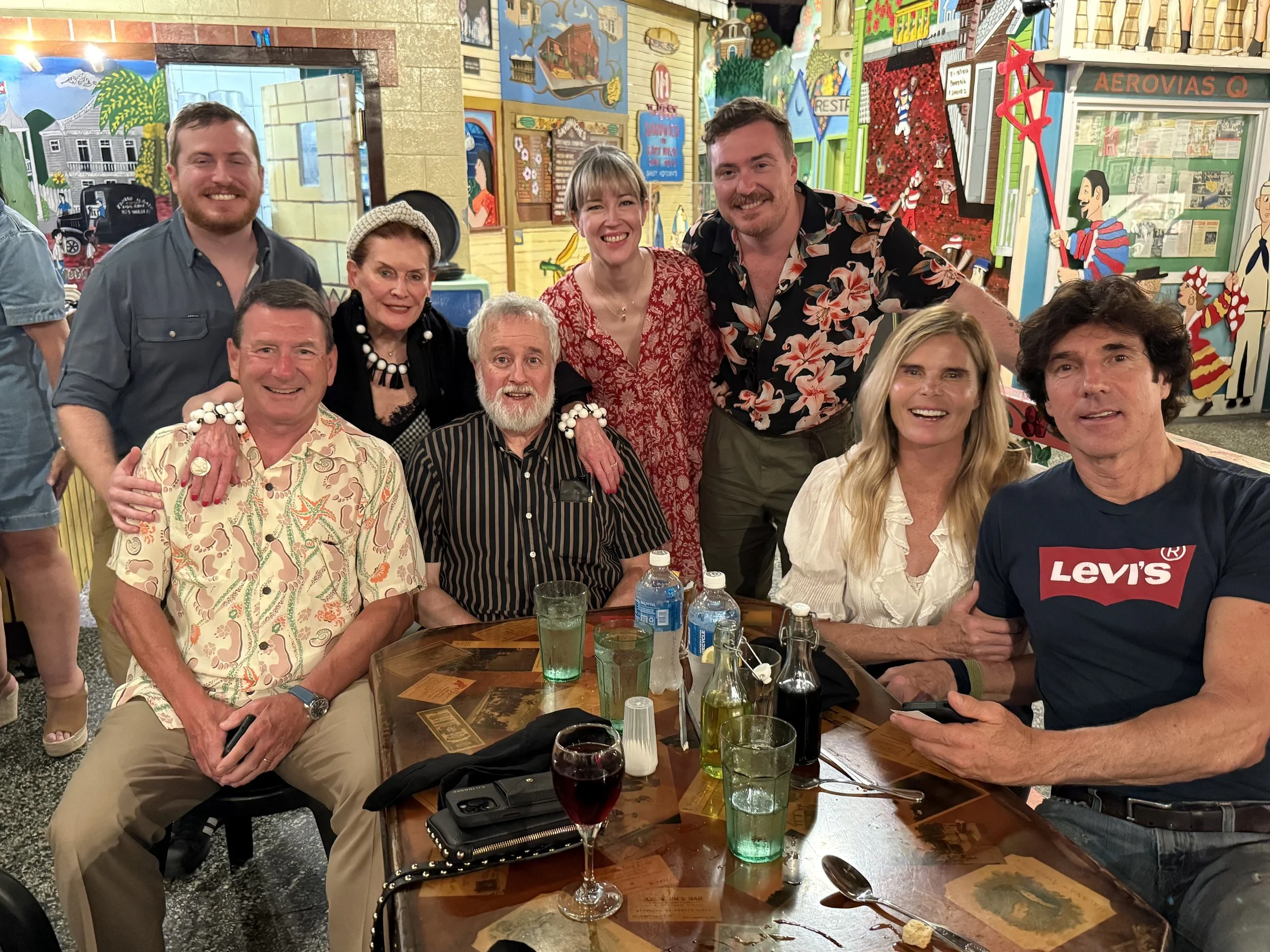 Group of eight people gathered around a dinner table in a colorful, decorated restaurant. Smiling and enjoying each other's company. The background features vibrant, artistic murals.