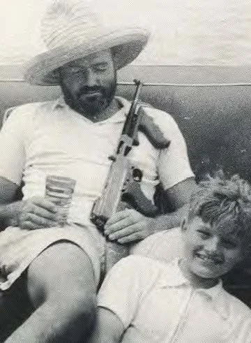 Man wearing a large sun hat, holding a rifle, sitting outdoors with a young boy, both smiling.