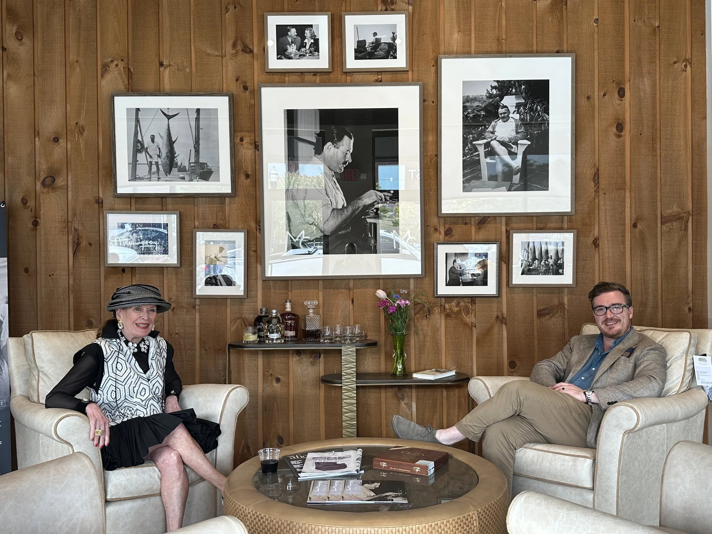 Two people, an older woman and a younger man, sitting on beige armchairs in front of a wooden wall with framed black-and-white photographs. The woman is wearing a hat, jewelry, and a patterned top, and the man is wearing glasses, a blazer, and khakis