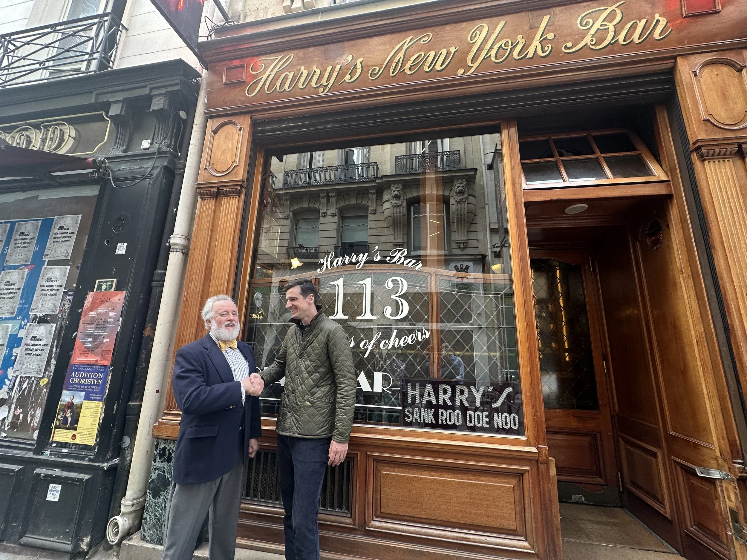 Two men shaking hands outside a bar named Harry's New York Bar. The bar has a wooden facade with gold lettering, and through the window, the bar's interior and some text on the window are visible. One man is older with a beard, wearing a dark suit an