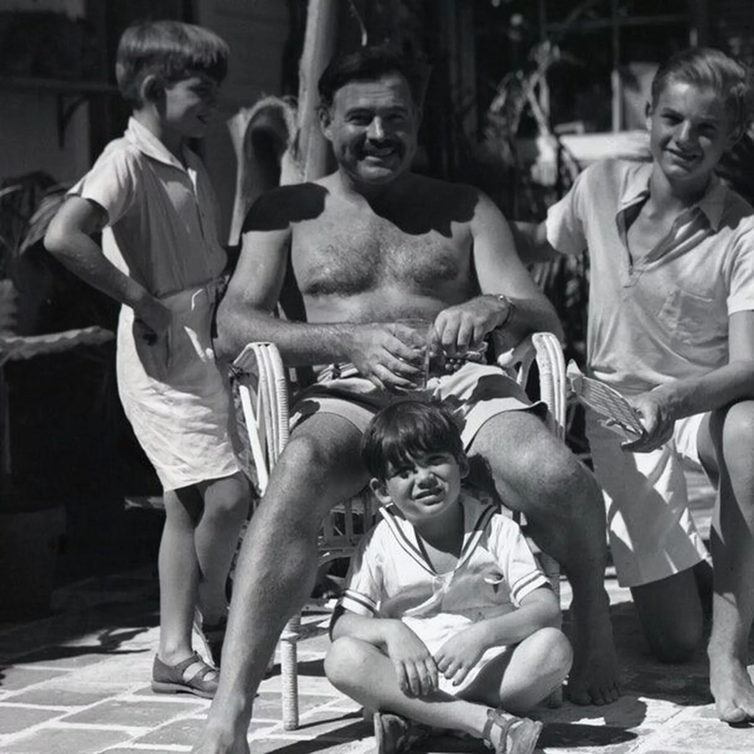 A black and white photo of a man sitting in a chair outdoors, flanked by two women, with a young boy sitting on the ground in front of him. The man is shirtless, smiling, and holding an object in his hands. The women are smiling and holding books, and the boy is squatting, looking at the camera.