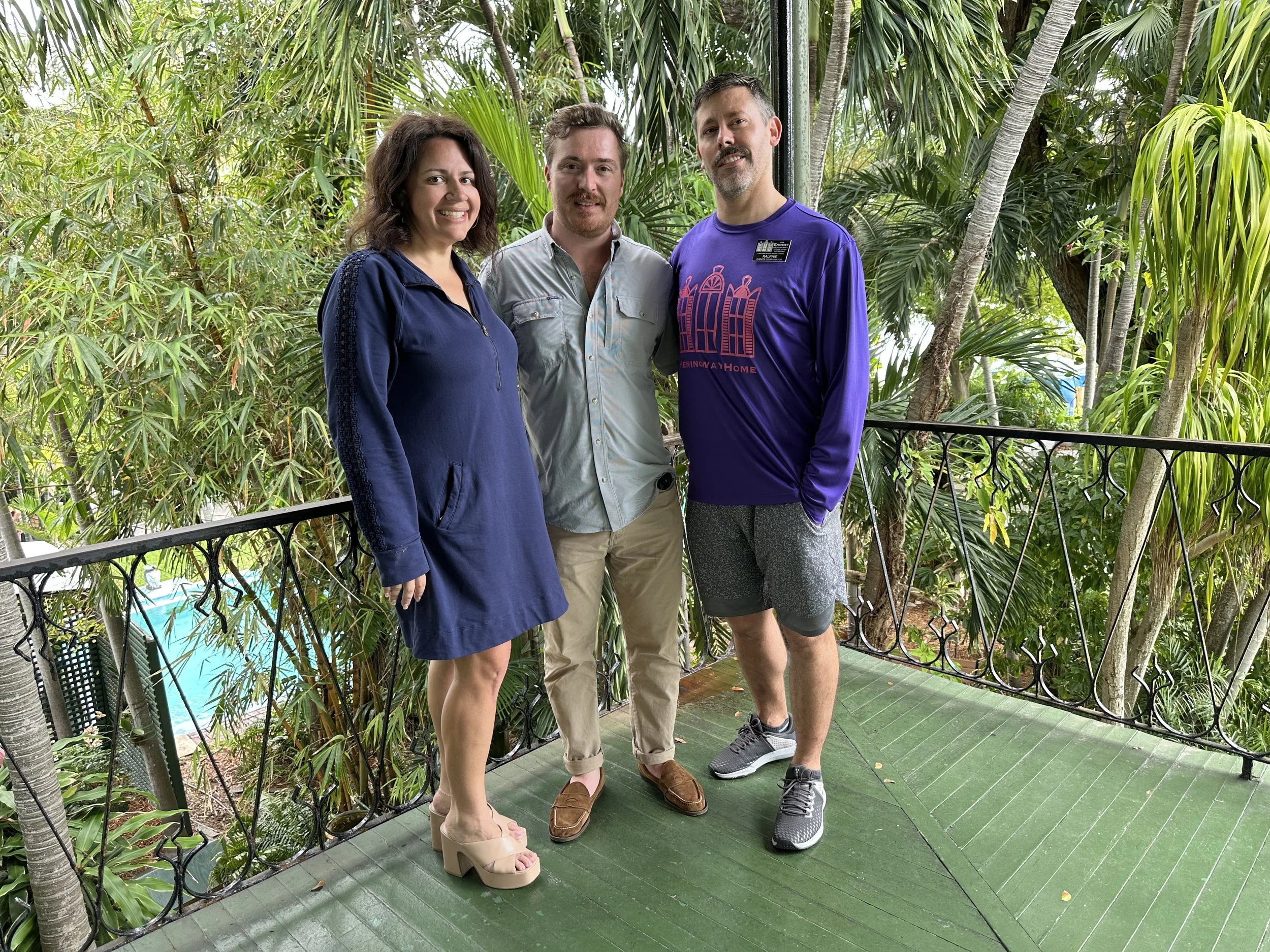 Three people standing on a green deck surrounded by lush green tropical plants, smiling at the camera.
