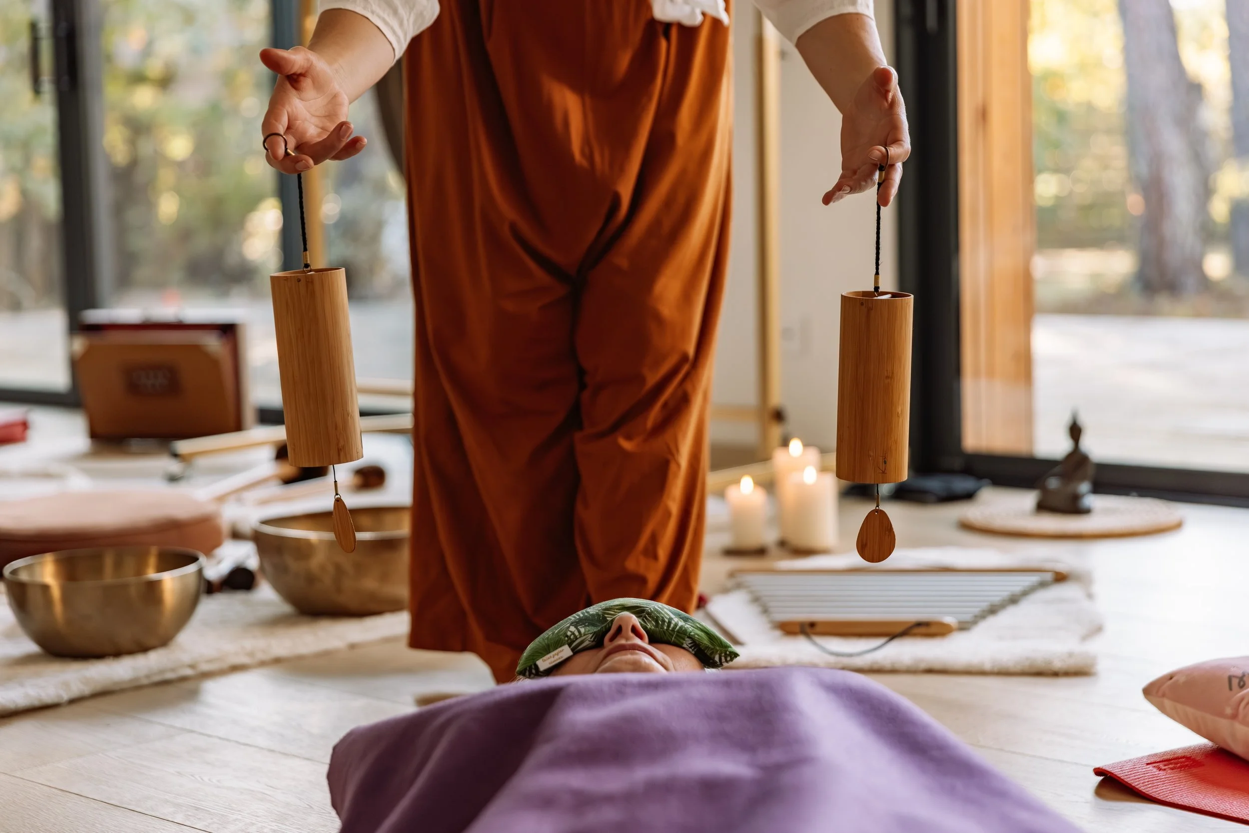 Une personne allongée sur un tapis lors d'une séance de relaxation ou de thérapie, avec une autre personne tenant deux bols en bois suspendus, dans une pièce lumineuse avec des bougies et des objets de méditation.