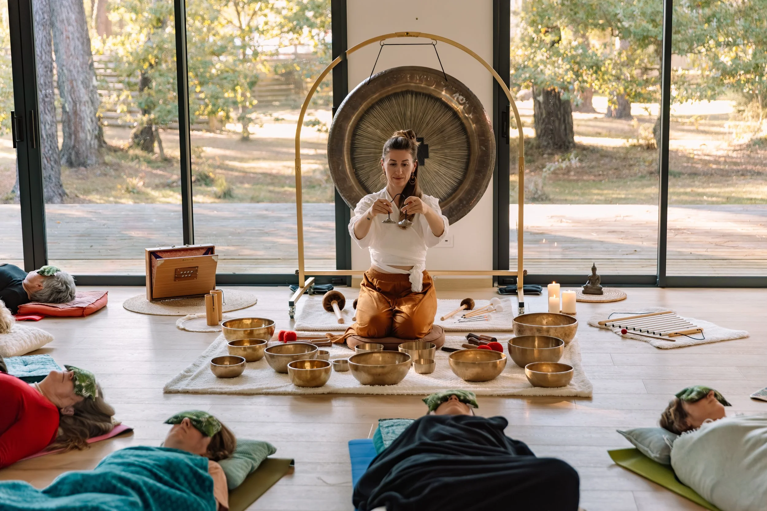 Une femme pratiquant une séance de relaxation ou de méditation dans une salle lumineuse avec de grandes fenêtres. Autour d'elle, plusieurs personnes allongées sur des tapis, portant des masques pour les yeux. Devant elle, des bols chantants en métal et d'autres instruments de musique. La scène évoque une ambiance zen et apaisante, avec un gong en arrière-plan.