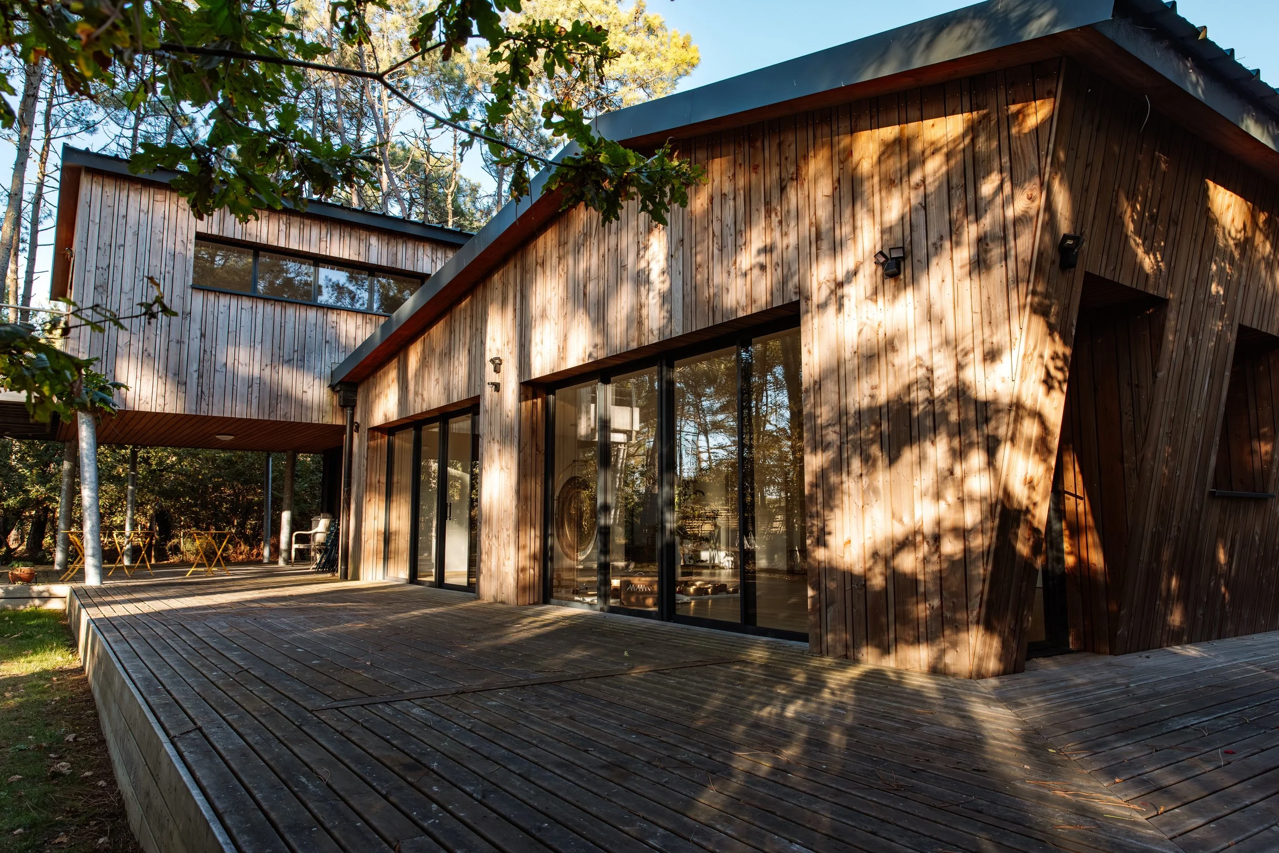 Maison en bois moderne avec une terrasse en bois, entourée d'arbres, en plein air par une journée ensoleillée.
