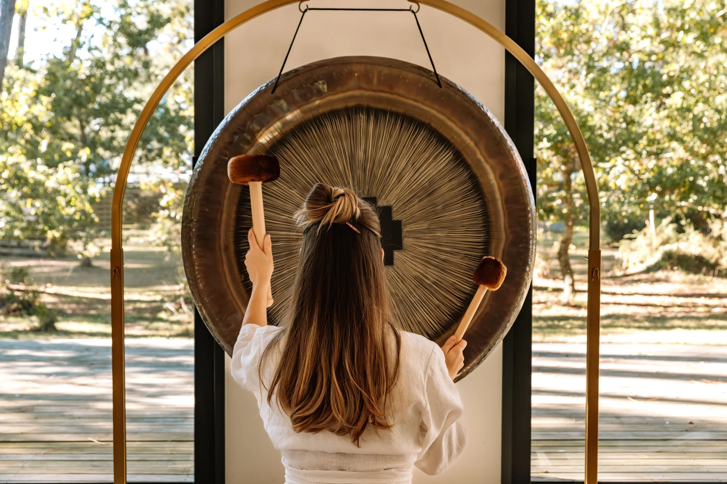 Une femme joue du gong en extérieur, vue de dos, avec la nature en arrière-plan.