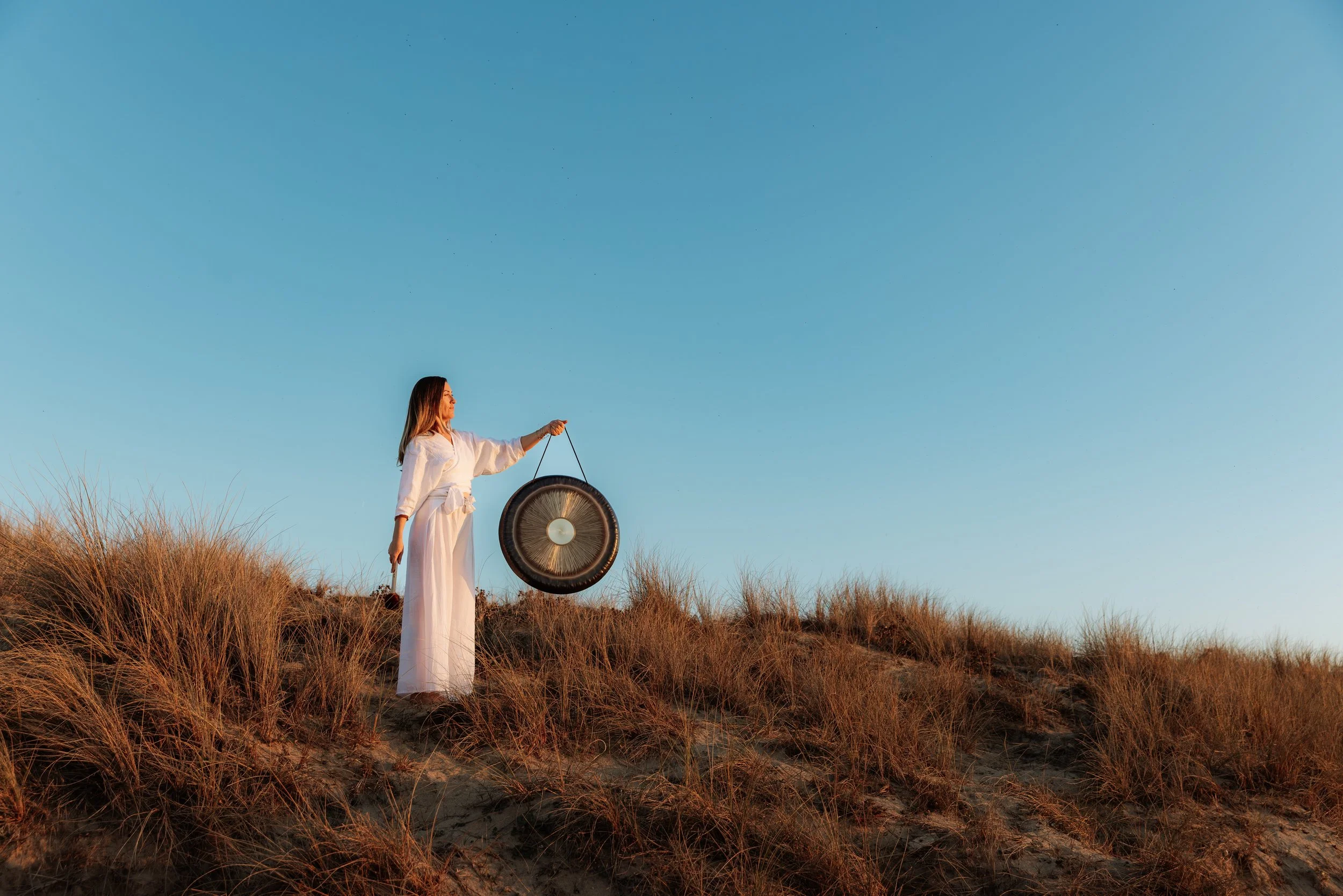Femme en robe blanche portant un gong en pleine nature lors du coucher du soleil