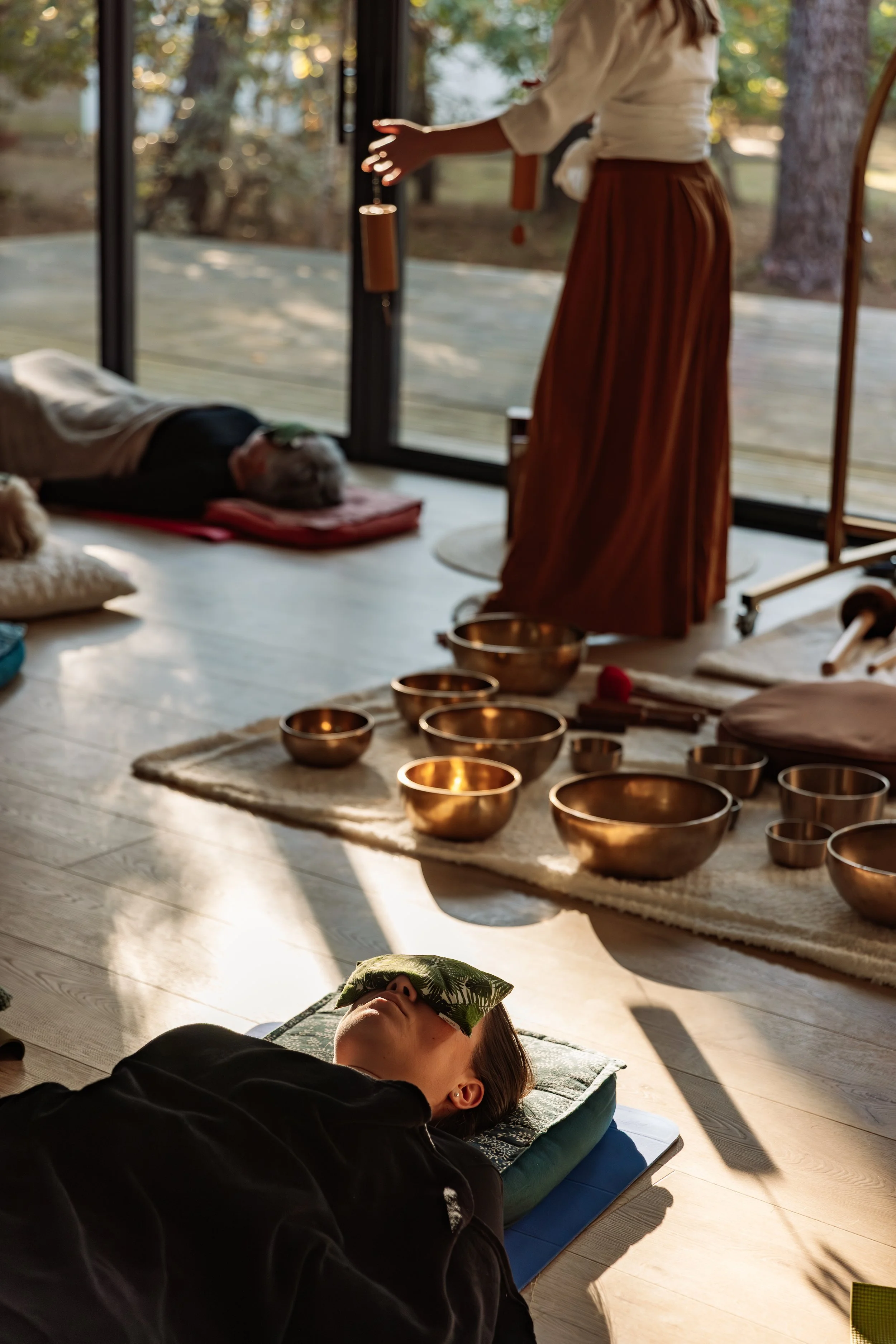 Deux personnes pratiquant une séance de relaxation ou de méditation, une allongée sur le sol avec un oreiller et l'autre à genoux, entourées de bols tibétains dans une pièce avec vue sur la nature.