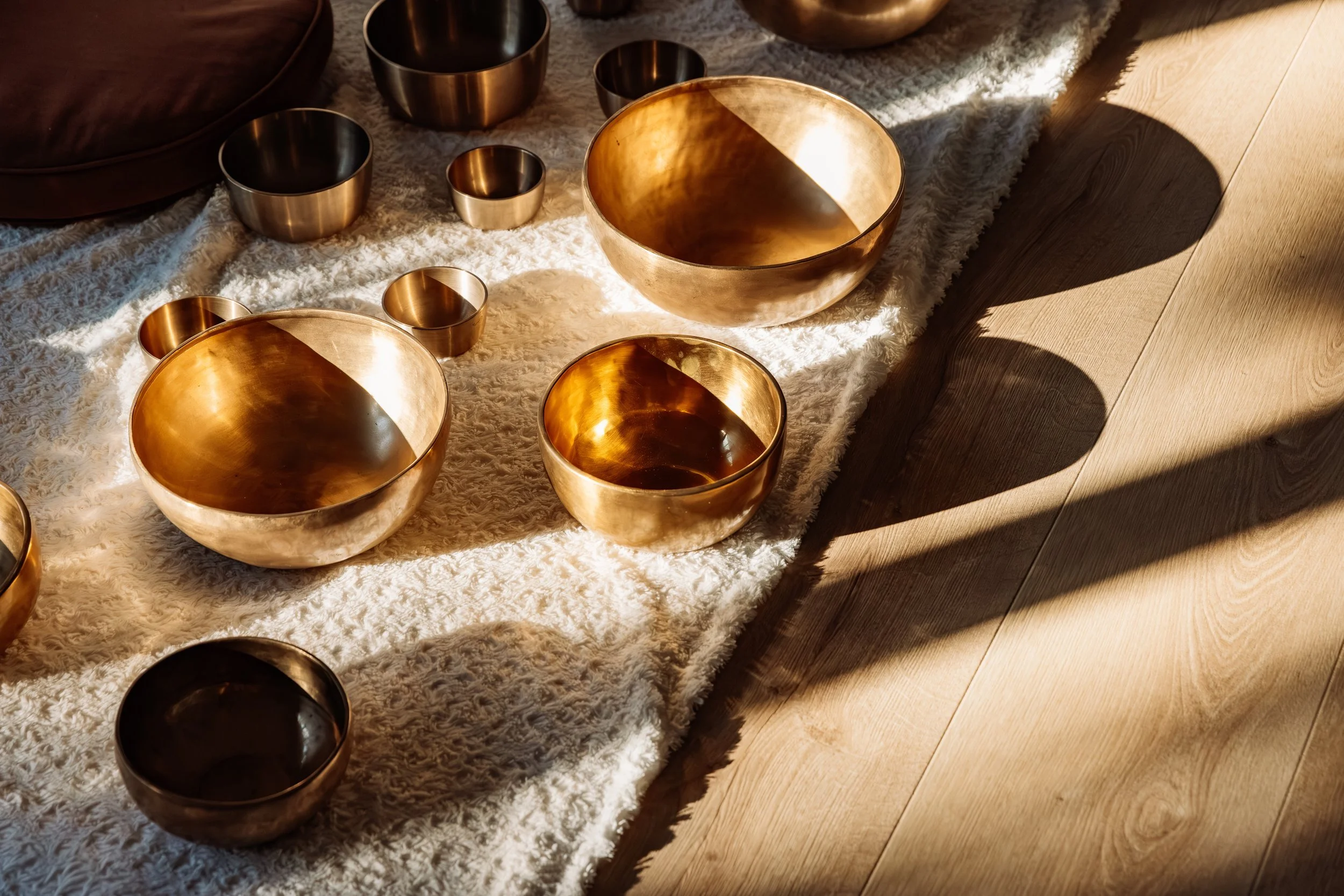 Une collection de bols en métal doré de différentes tailles posés sur un tapis blanc, avec des ombres projetées sur un sol en bois.