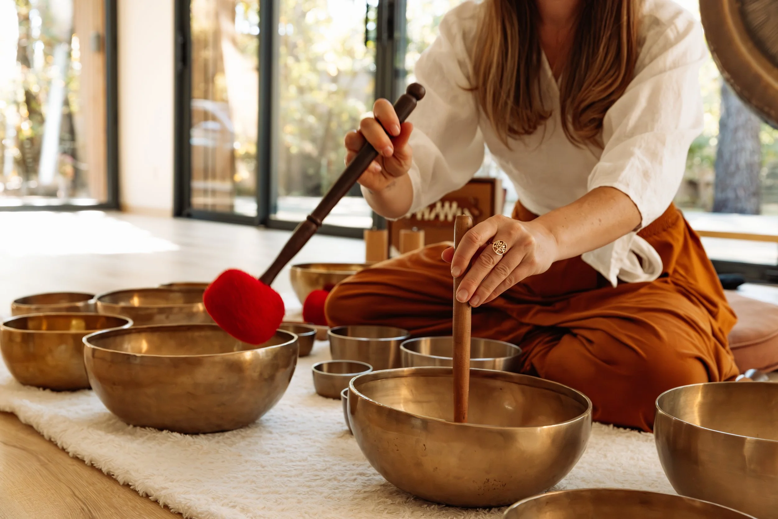 Une personne assise par terre joue des bols chantants en métal avec un maillet. La personne porte une robe orange et un vêtement blanc, et il y a plusieurs bols disposés sur un tapis blanc devant elle. La scène se déroule dans une pièce lumineuse avec de grandes fenêtres et des arbres visibles à l'extérieur.