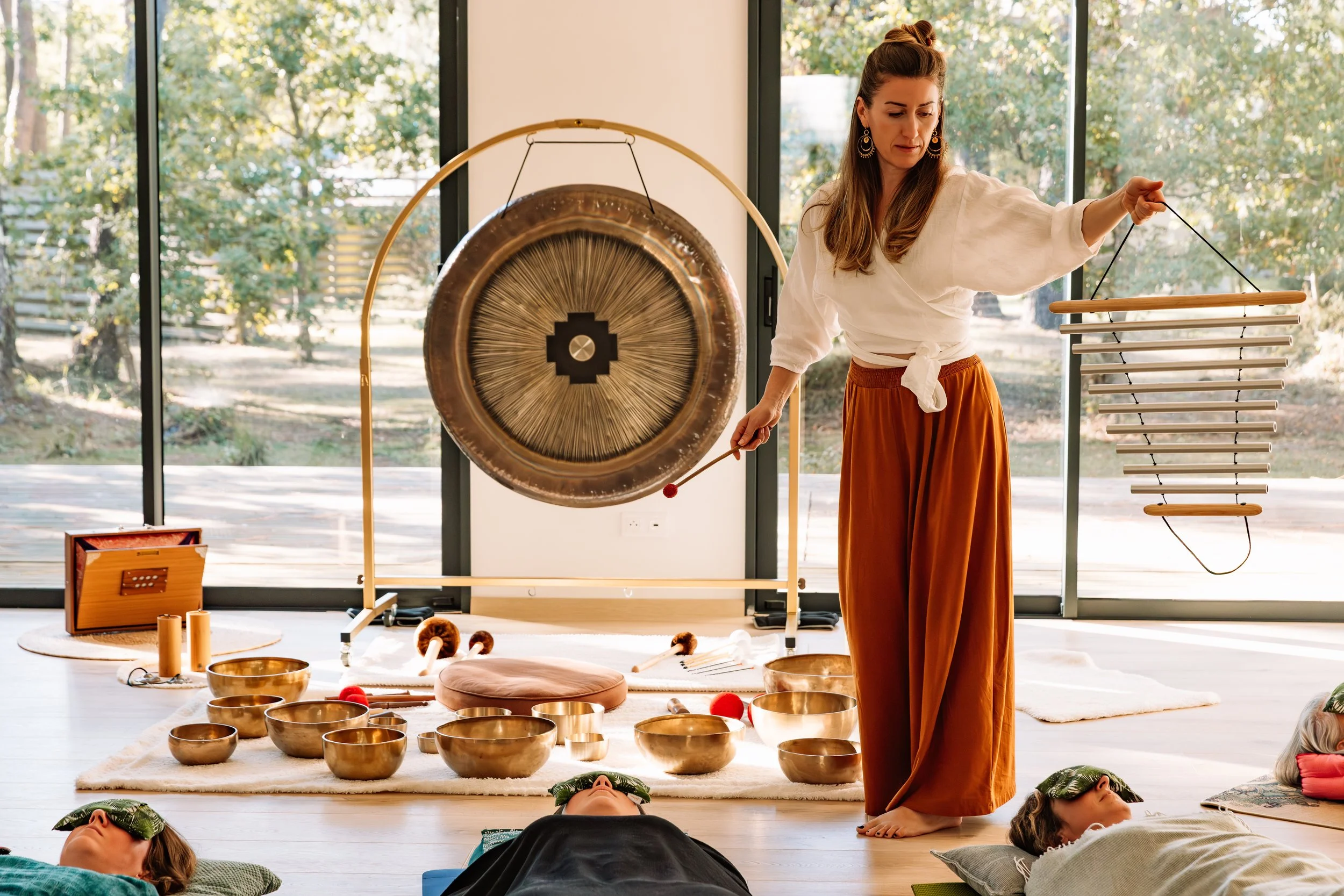 Une femme est en train de jouer du gong dans une salle lumineuse avec des fenêtres derrière lui, et plusieurs personnes sont allongées sur des tapis en train de méditer ou de se détendre.