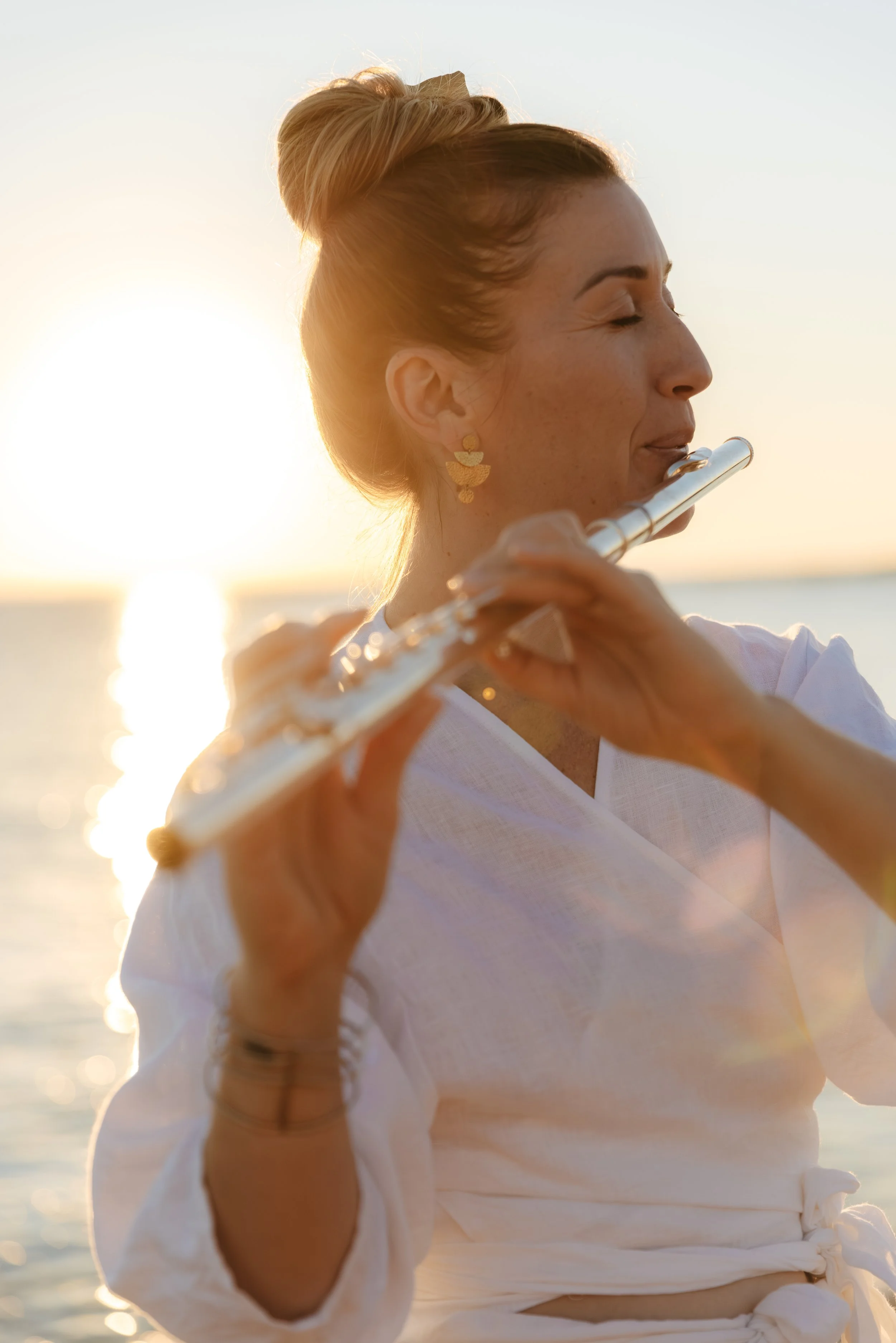 Une femme jouant de la flûte au coucher du soleil près de la mer, portant une tenue blanche et des boucles d'oreilles en forme de demi-lune.