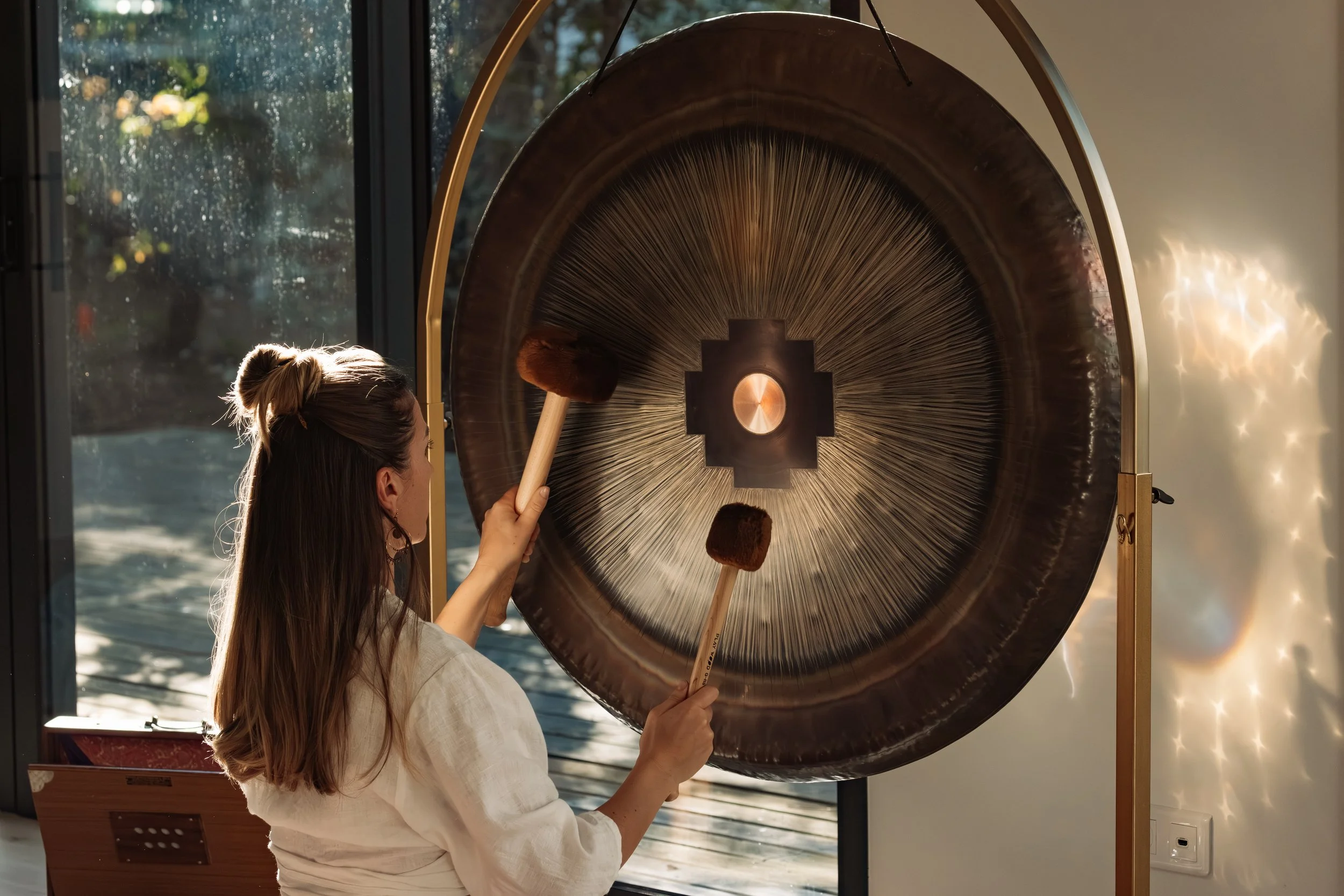 Une femme jouant du gomp à l'aide de deux énormes mallets en caoutchouc dans une pièce éclairée par la lumière naturelle, avec une grande cloche en métal devant elle.
