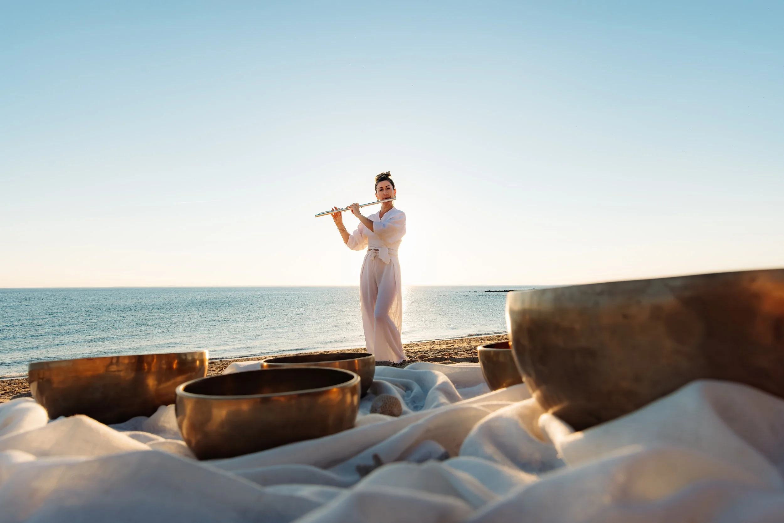 Une femme joue de la flûte sur la plage lors du coucher de soleil, avec des bols en bronze posés sur un drap blanc devant elle.
