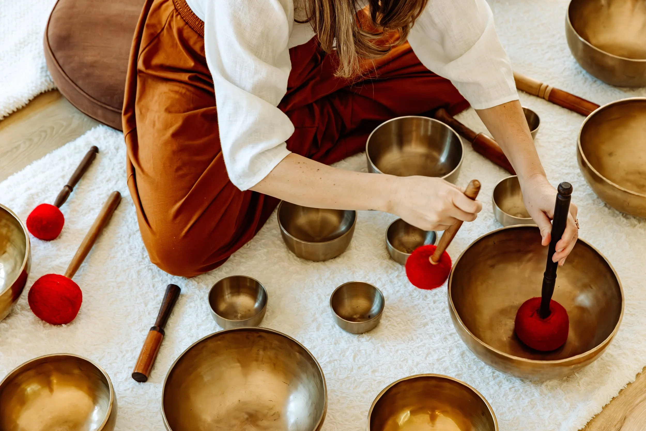 Une personne joue du bol chantant en métal, entourée d'autres bols et bâtons de mallet, sur un tapis moelleux.