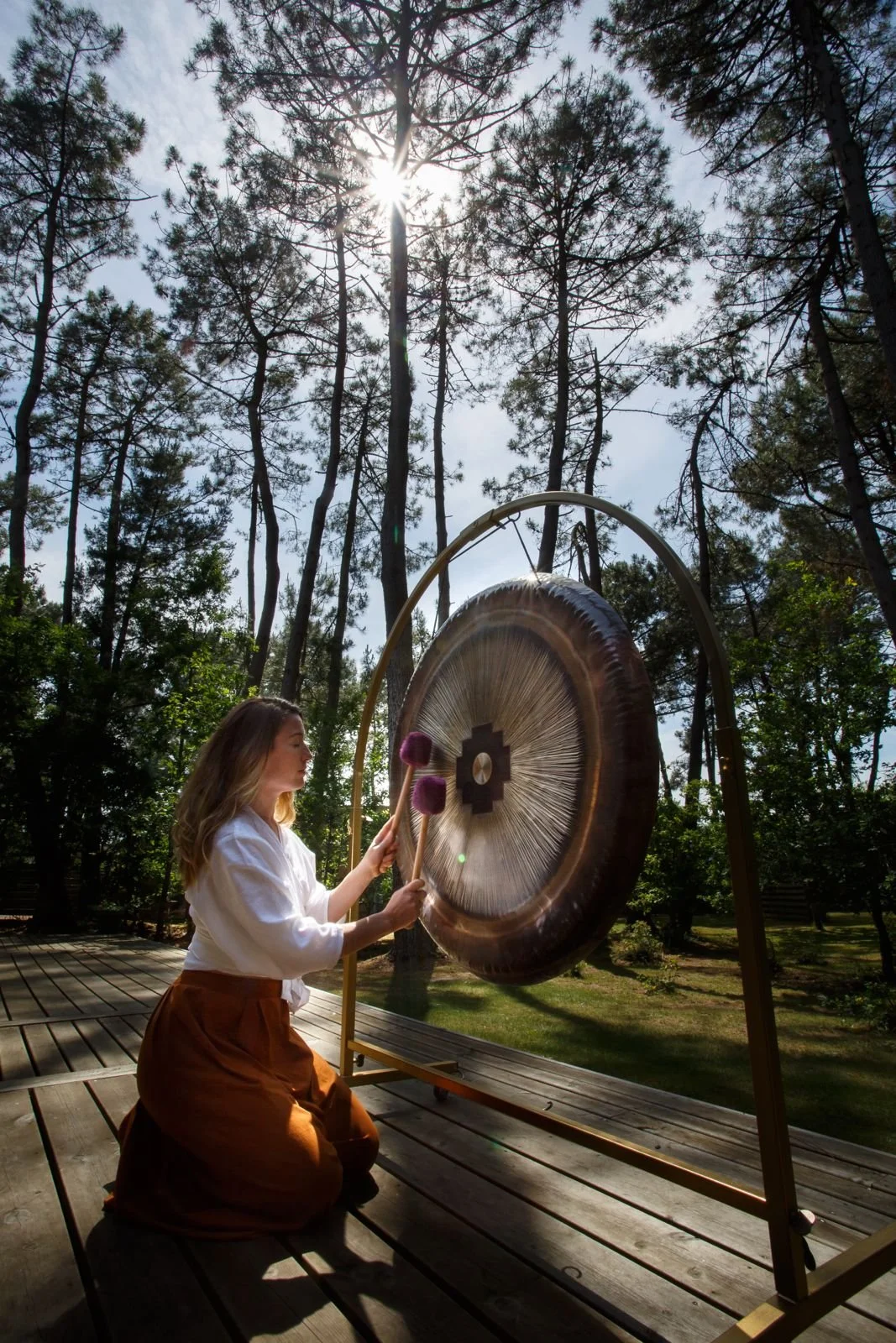 Jeune femme jouant du gong en plein air dans une forêt, avec le soleil brillant à travers les arbres.