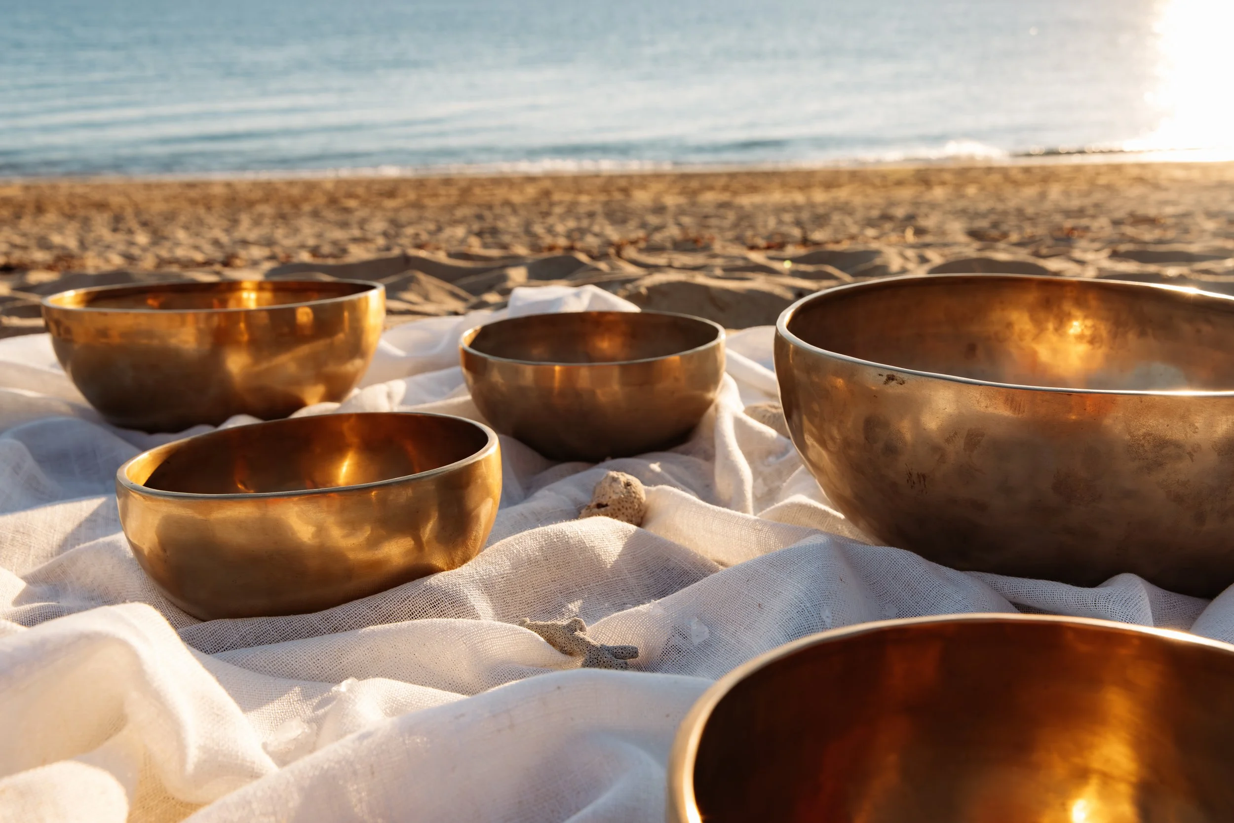 Cinq bols en métal doré placés sur des draps blancs sur la plage, avec la mer et le soleil couchant en arrière-plan.