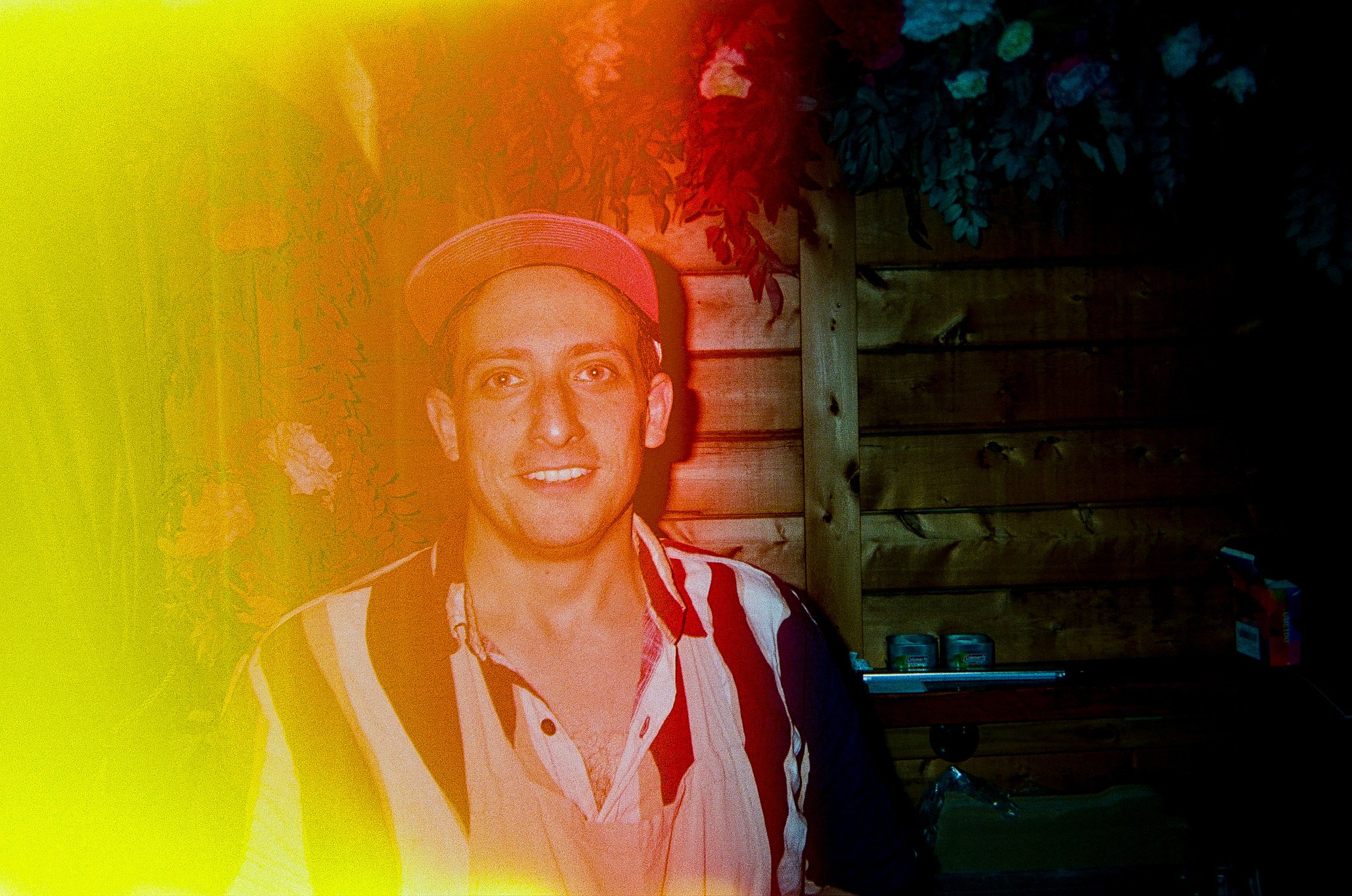 A man wearing a light-colored baseball cap and a striped shirt, smiling, standing outdoors at night with a wooden fence and foliage in the background, illuminated by warm lighting.