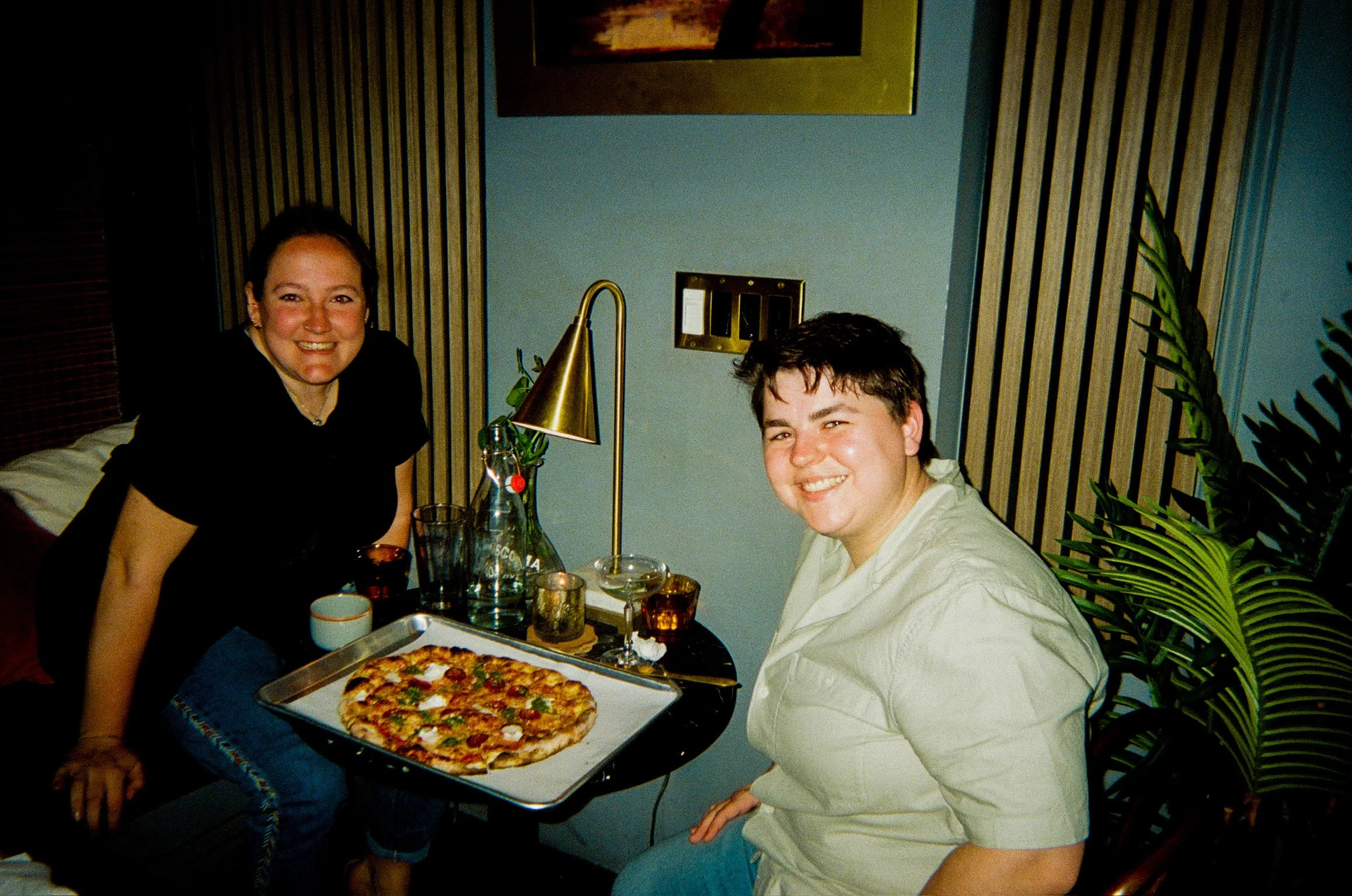 Two smiling people, a woman and a man, sitting at a table with a pizza and drinks in a cozy indoor setting with wooden paneling and plants.