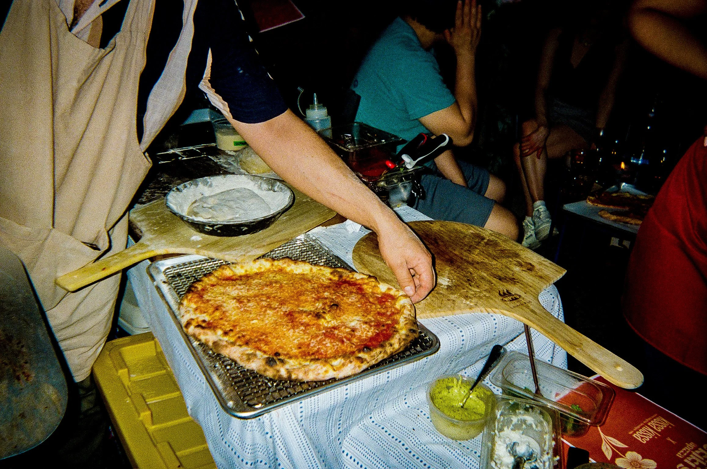 A person preparing a pizza at a food stall or restaurant, with a pizza on a tray, a cutting board, and various ingredients and utensils on the counter.