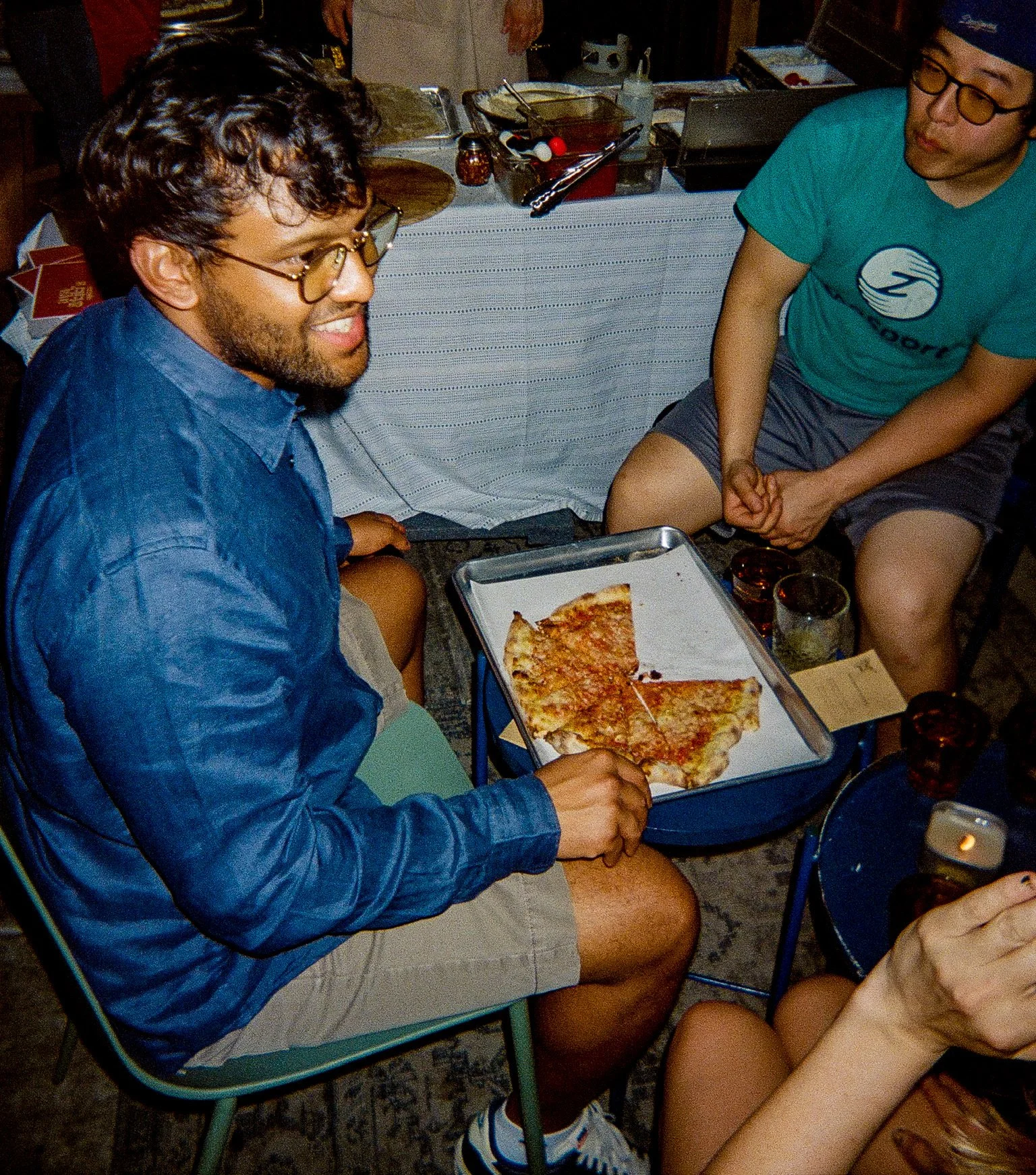 Two people sitting at a table with pizza, talking and laughing, with drinks nearby in a casual setting.