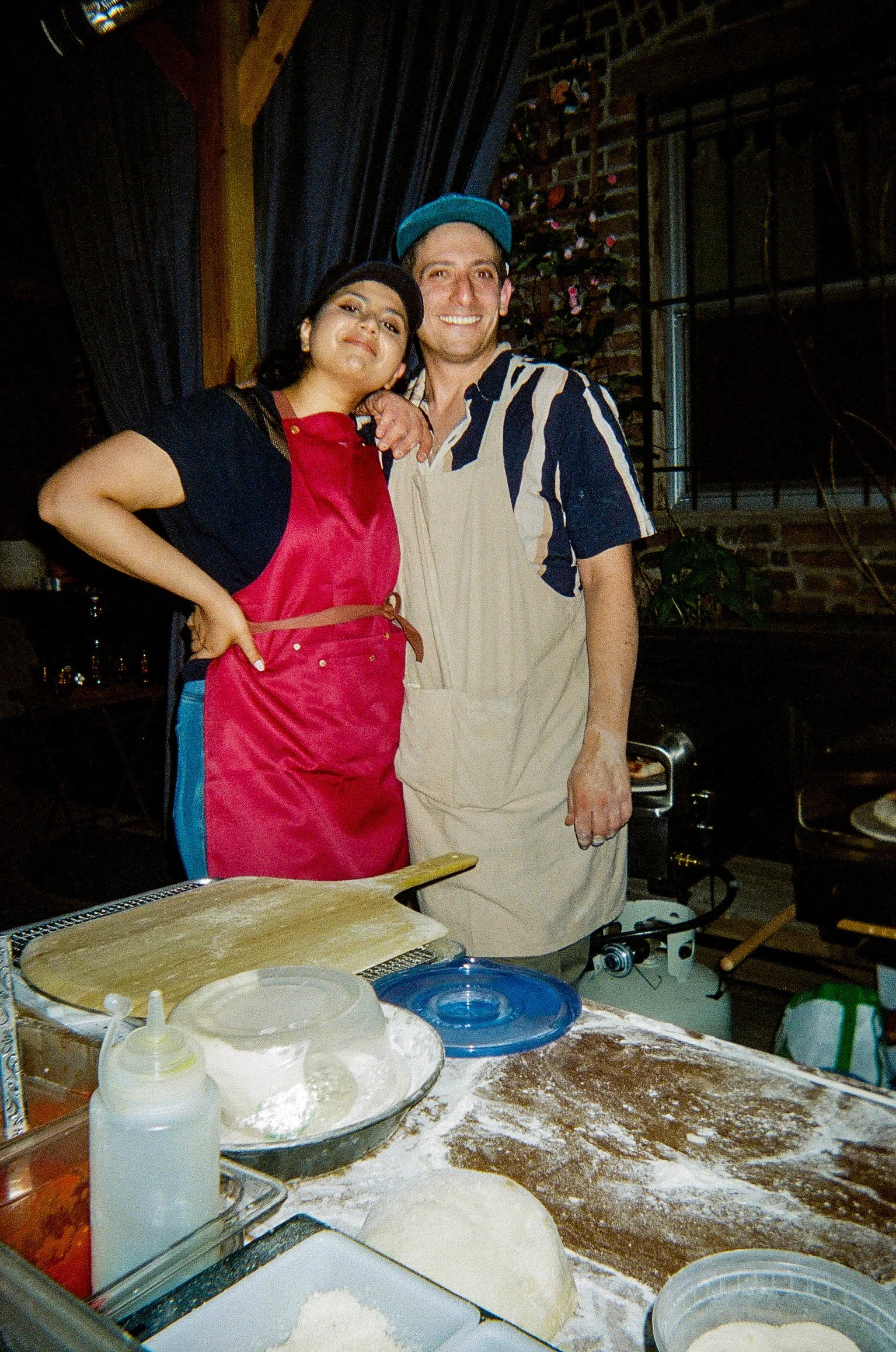 Two people, a woman in a red apron and a man in a beige apron, standing together in a kitchen or bakery setting, smiling, with dough, baking tools, and ingredients on the table in front of them.