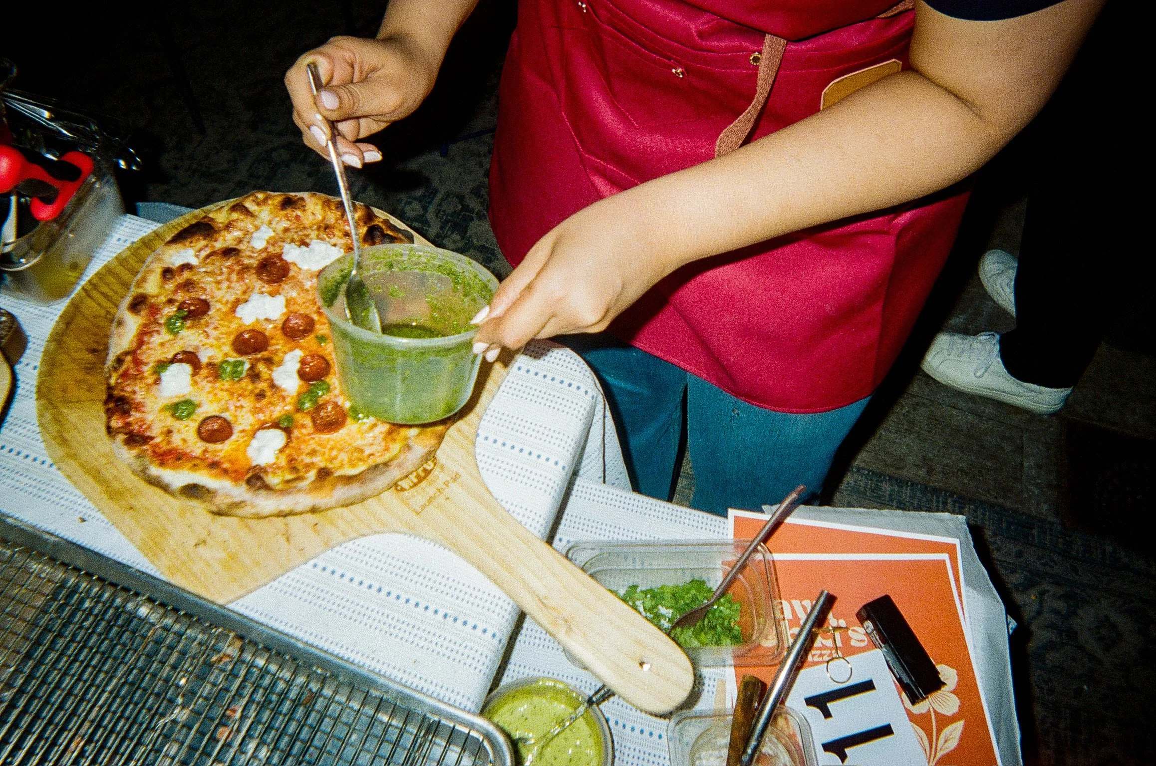 Person serving pizza with pesto, tomato sauce, burrata, and pepperoni on a  dining table with salads, condiments, and menu items.