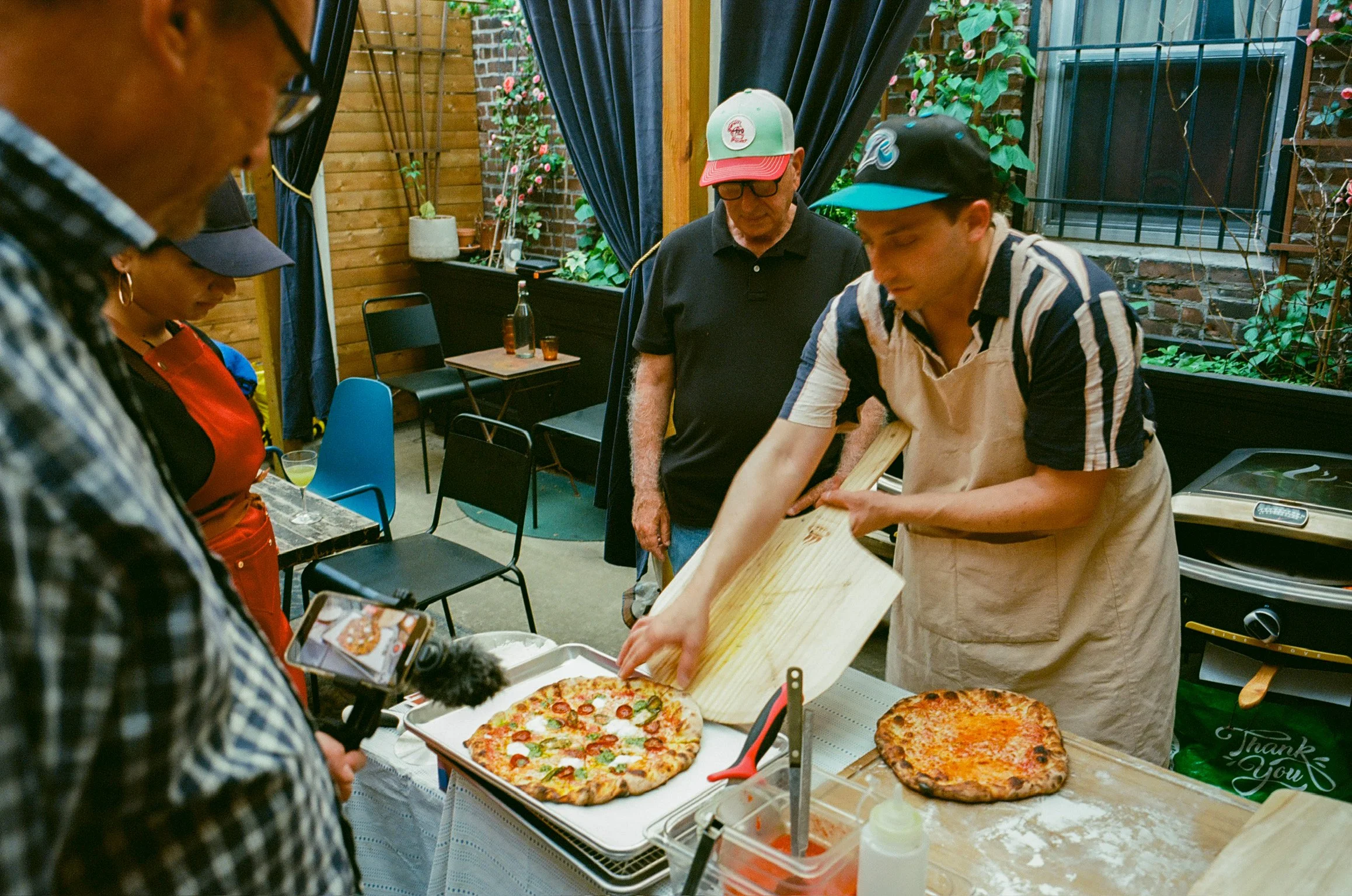 A man in a striped shirt and apron serves a New Haven-style pizza at an outdoor gathering, while others watch, with a backdrop of a garden and a brick wall.