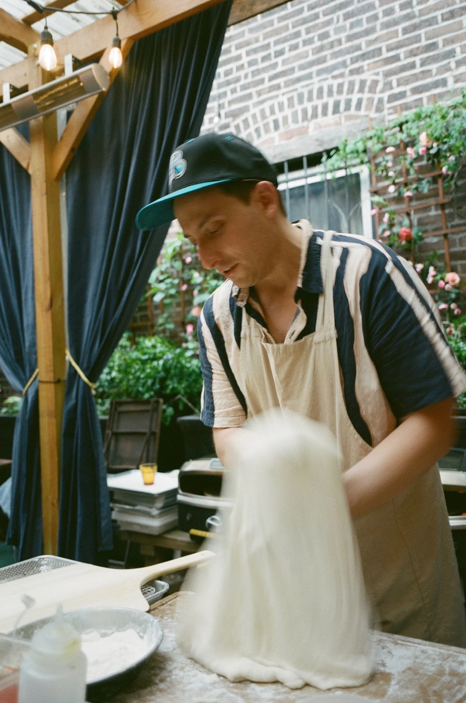 A man in a black cap and striped shirt stretching and folding dough on a wooden surface in a cozy outdoor kitchen with brick wall and greenery.