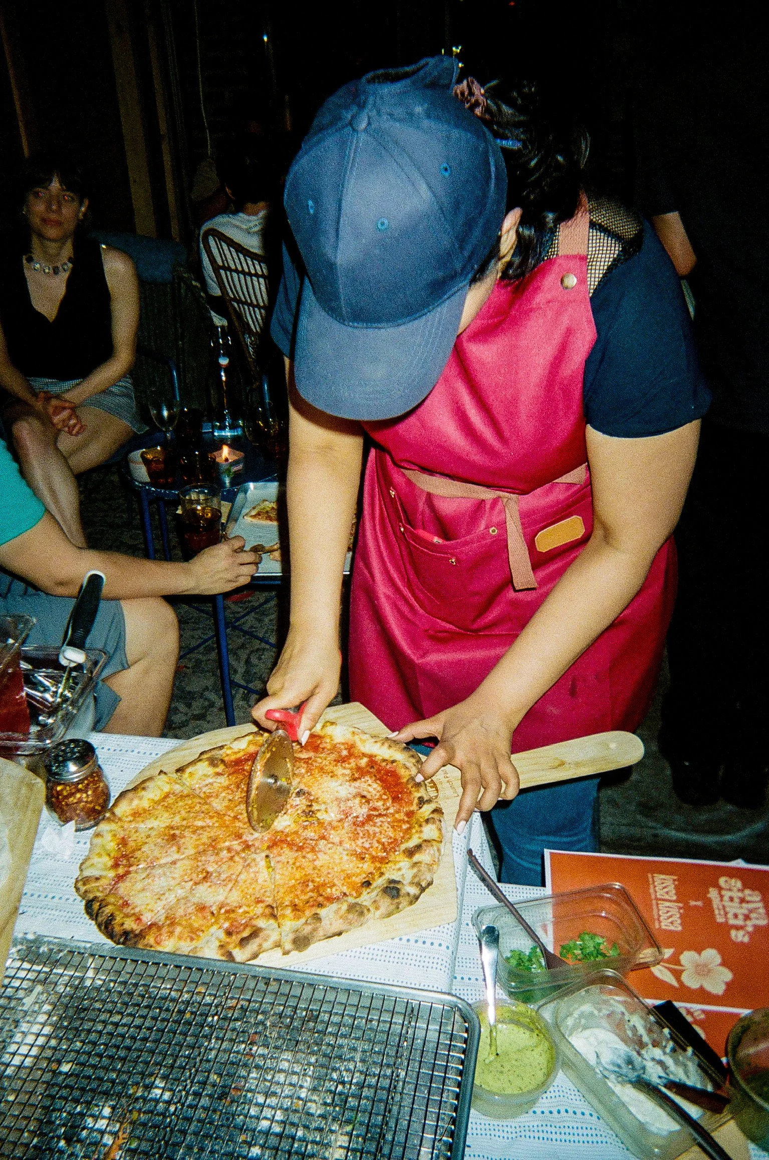 Person slicing a large pizza at a restaurant table with other seated guests nearby.