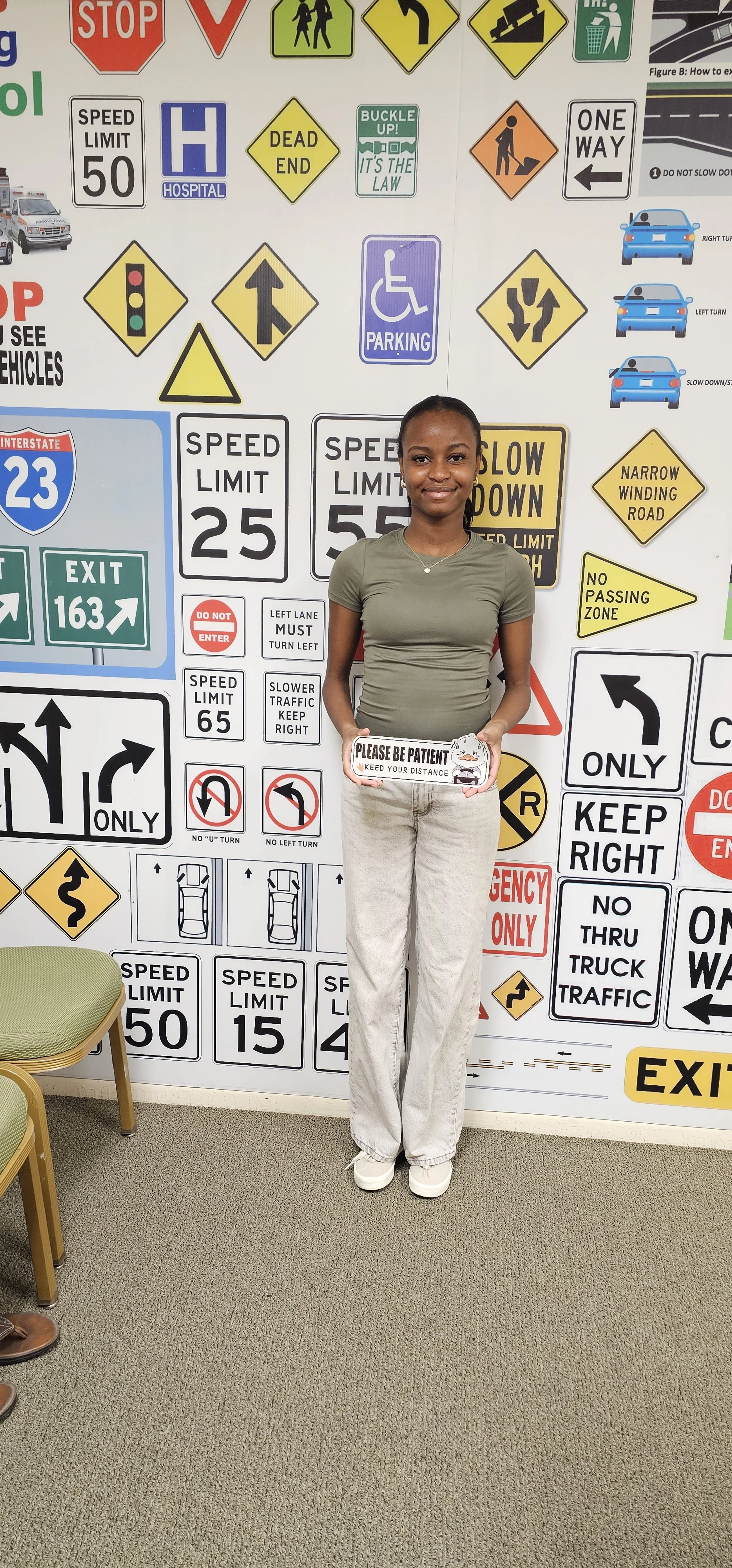 Young woman standing in front of a wall covered with traffic signs, holding a sign that says 'Please Be Patient Keep Your Distance' inside an office or classroom setting.