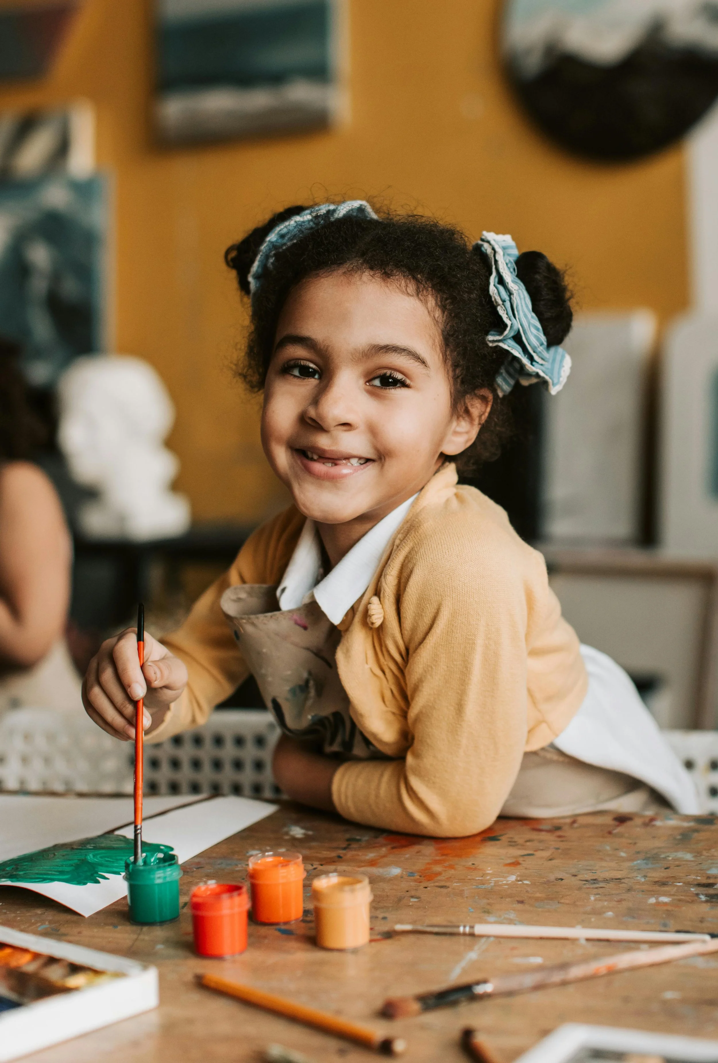A young girl with curly hair in pigtails with striped scrunchies, smiling while engaging in painting art in a classroom. She is using a paintbrush to apply green paint on a paper, surrounded by containers of paint and art supplies on a wooden table.
