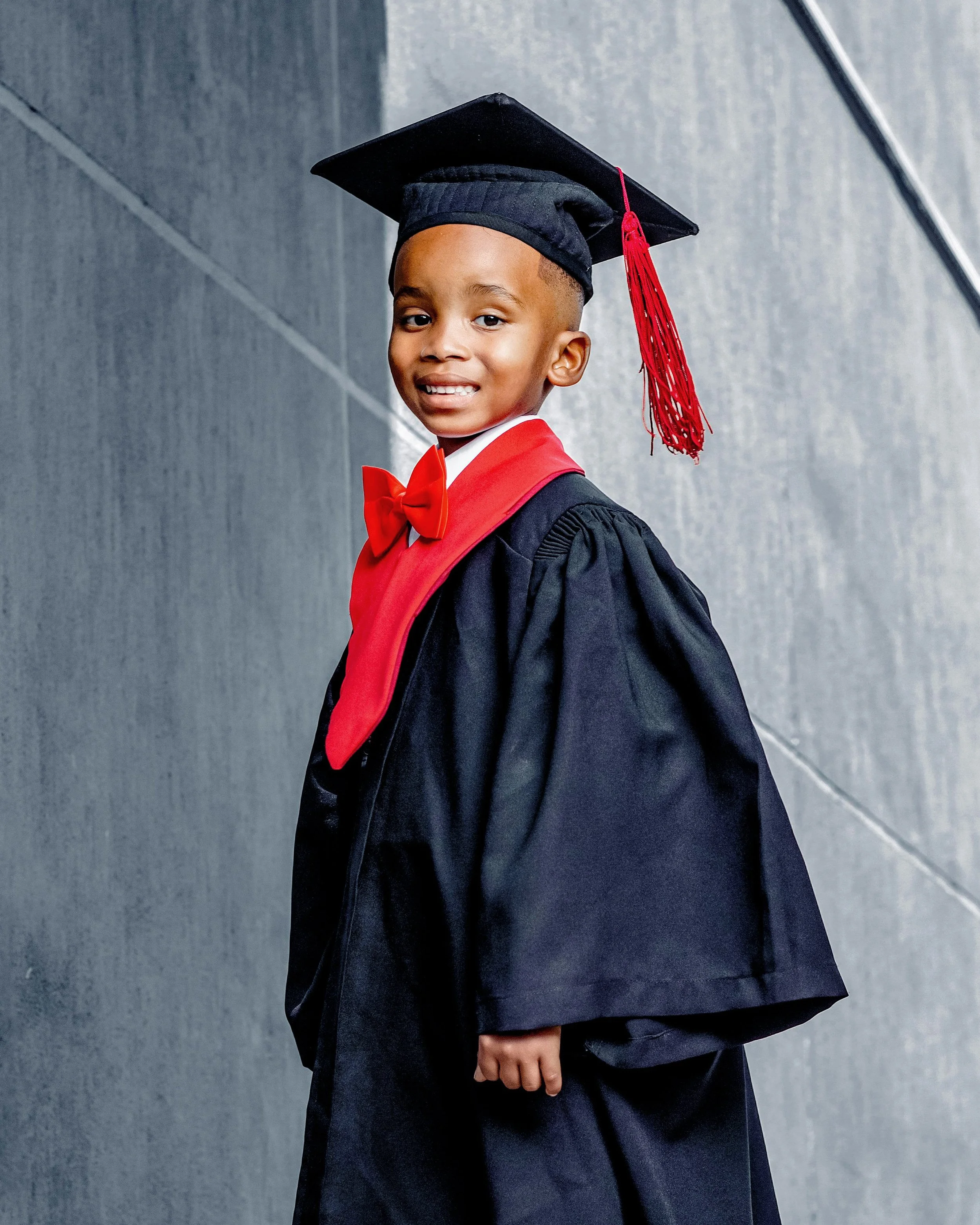 Portrait of a young boy in a graduation gown and cap, with a red bow tie, standing against a gray textured background.