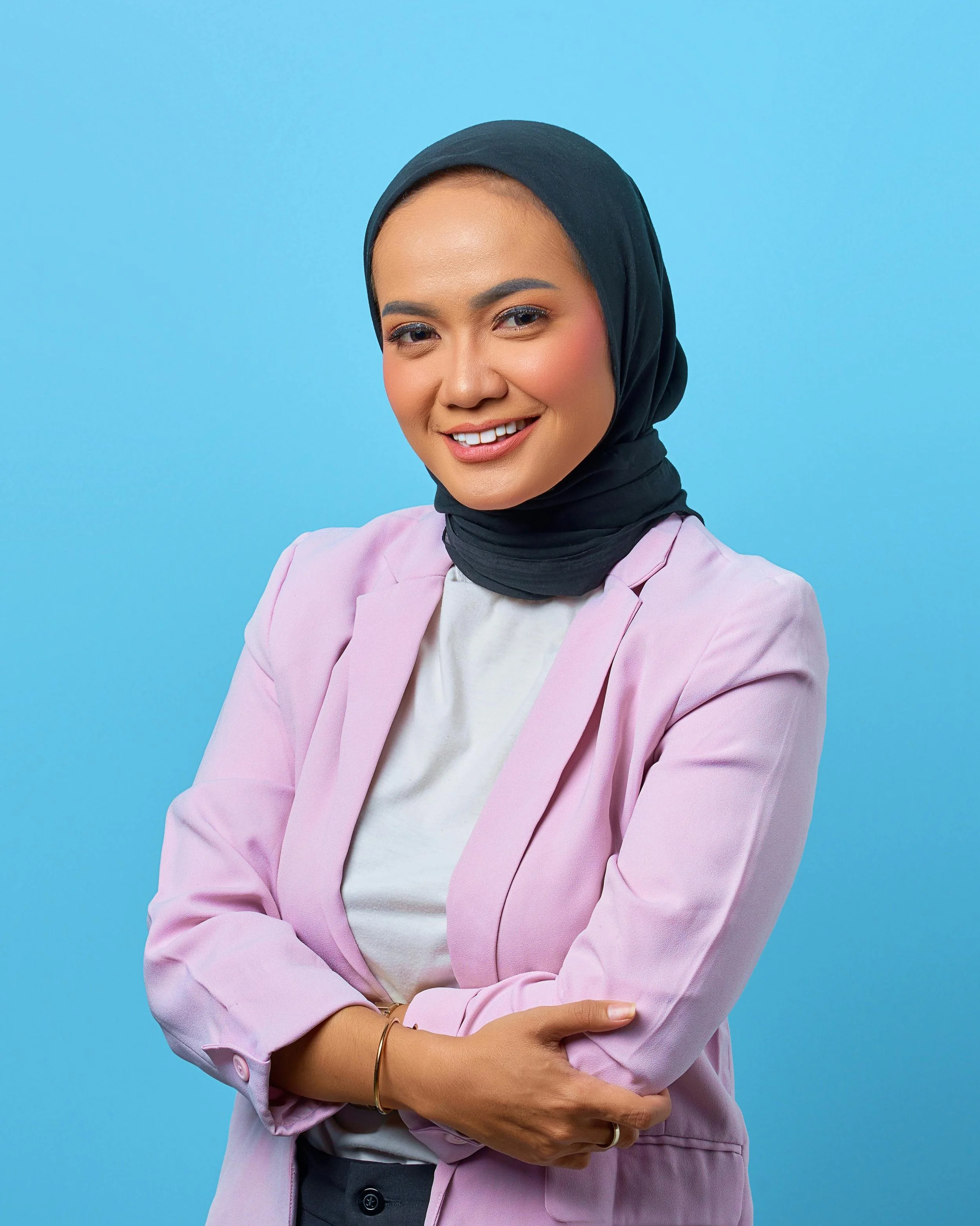 Close-up of a woman wearing a navy blue hijab, pink blazer, and white shirt against a light blue background. She is smiling and has her arms crossed.