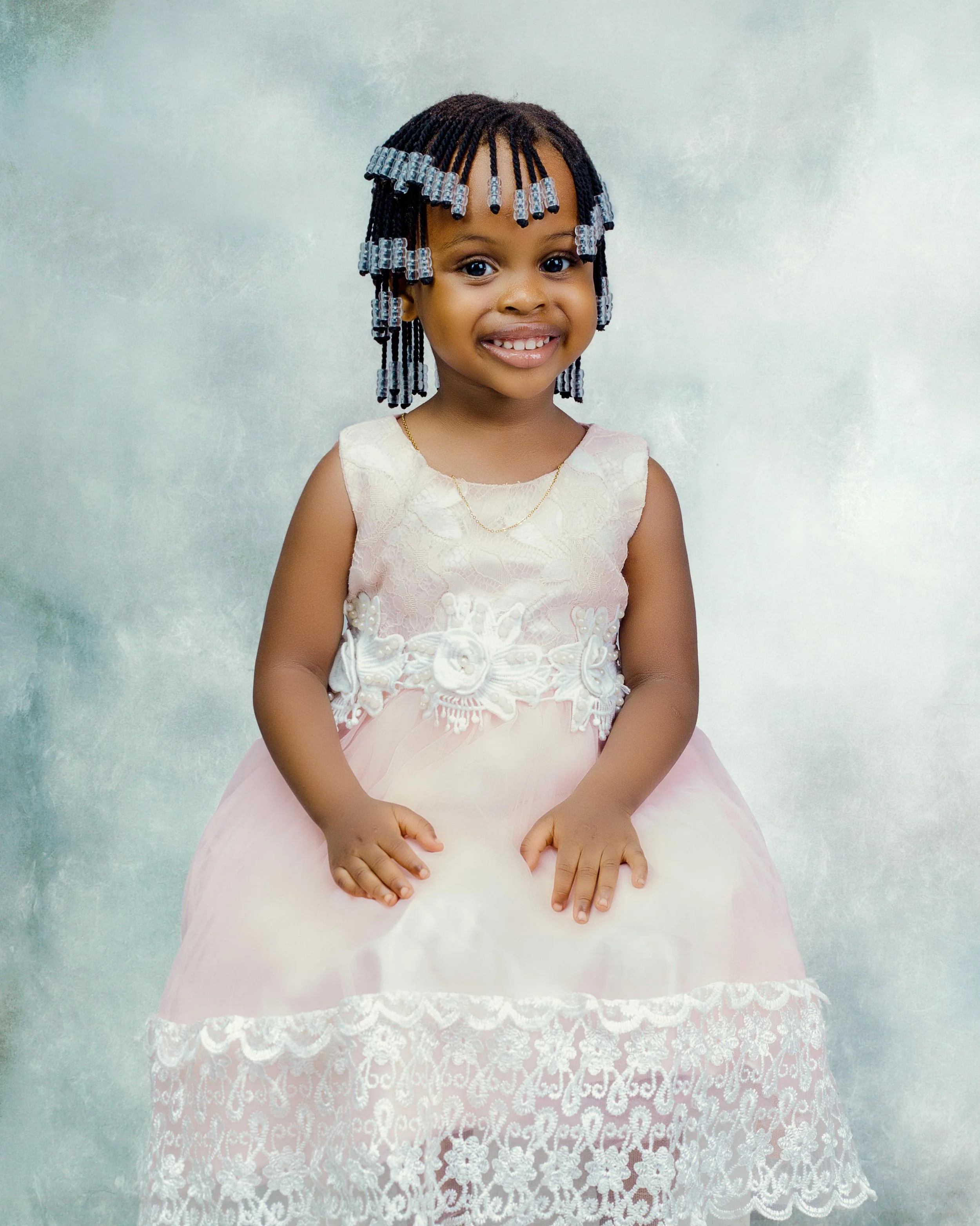 A young girl with braided hair decorated with transparent beads, smiling, wearing a sleeveless cream-colored dress with lace and floral embroidery, and a delicate gold chain necklace, standing in front of a light-colored textured background.