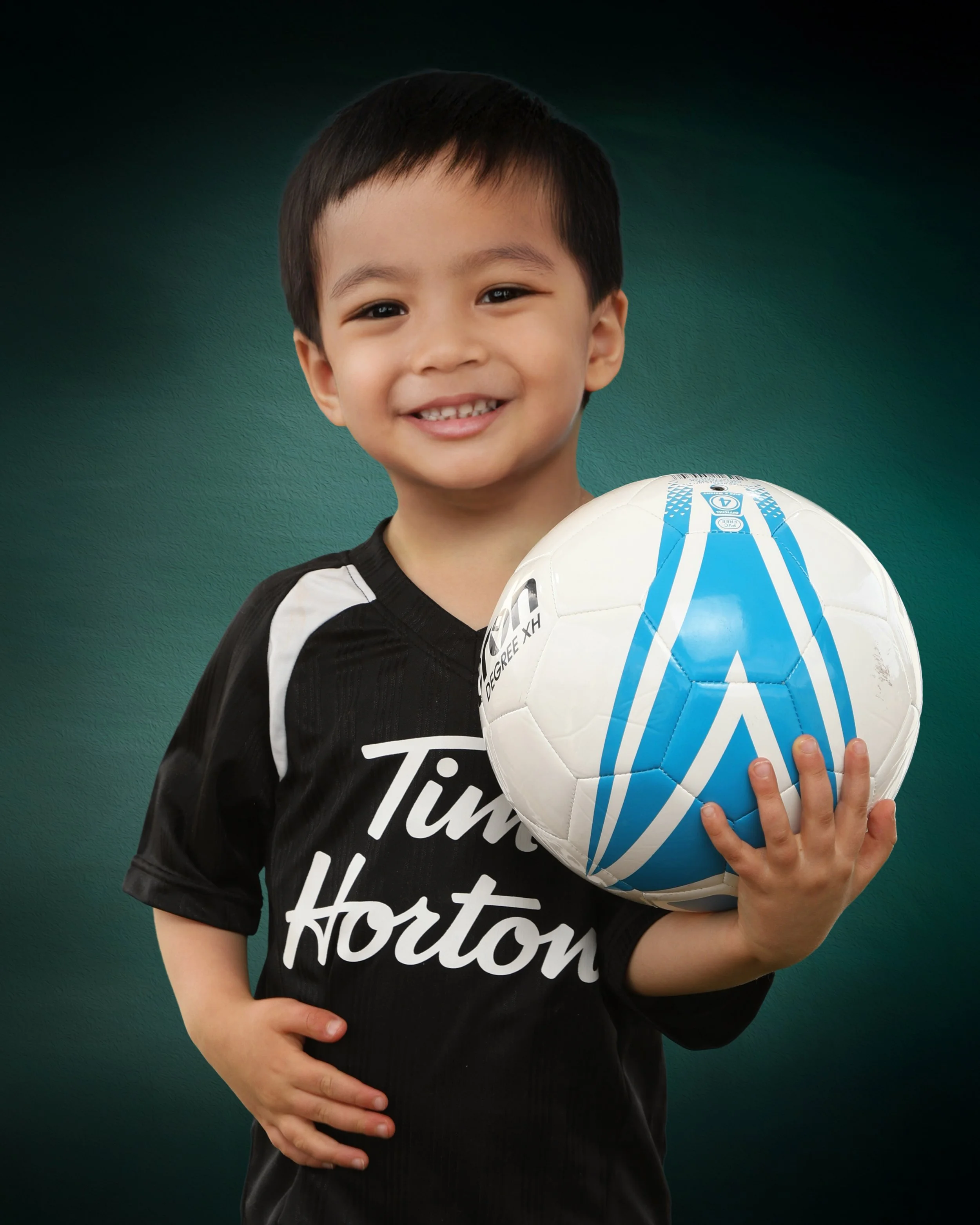 A young boy with short dark hair, smiling and holding a soccer ball in front of a dark green background. He is wearing a black and white sports jersey with the word 'Huston' visible on the front.