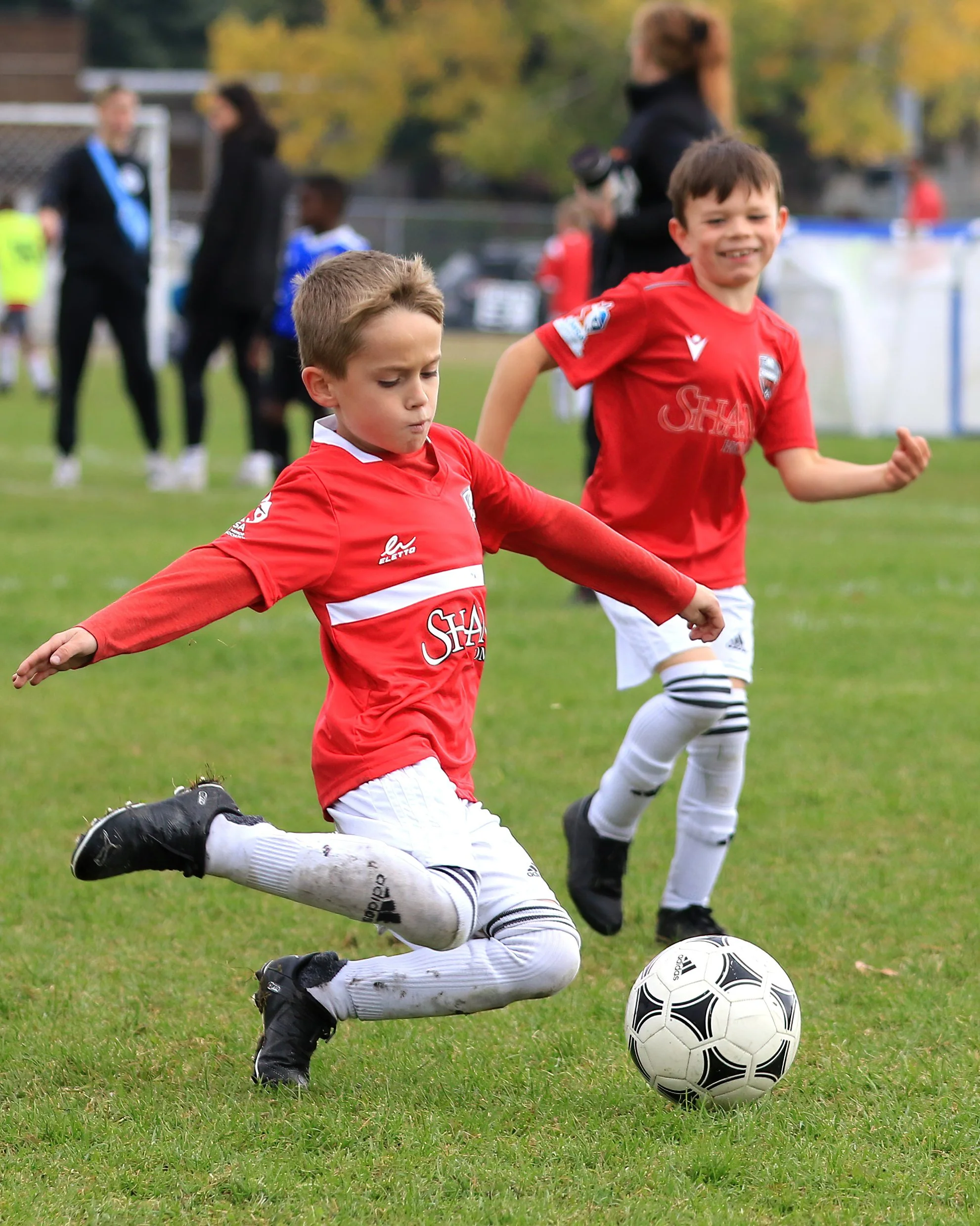 Two young boys in red soccer uniforms playing soccer on a grass field. One boy is kicking the ball with his right foot, and the second boy is smiling and running nearby. In the background, adults and children are visible, some watching and others wal