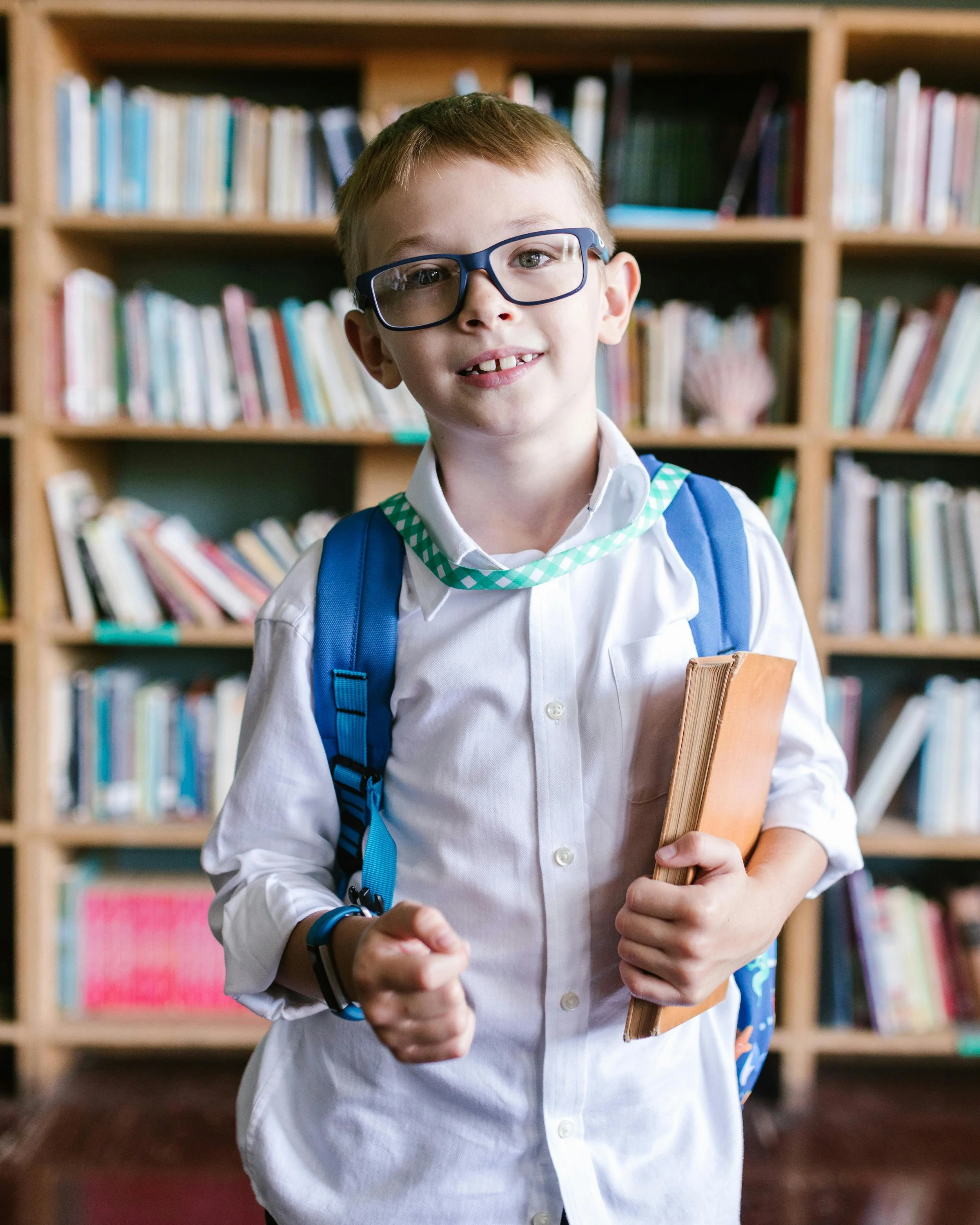 A young boy with glasses, a white shirt, and a backpack holding a book stands in a library with shelves filled with books in the background.