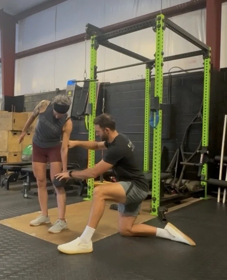 A man kneeling on the floor offering support and encouragement to a woman lifting her leg, in a gym with workout equipment and a green power rack.